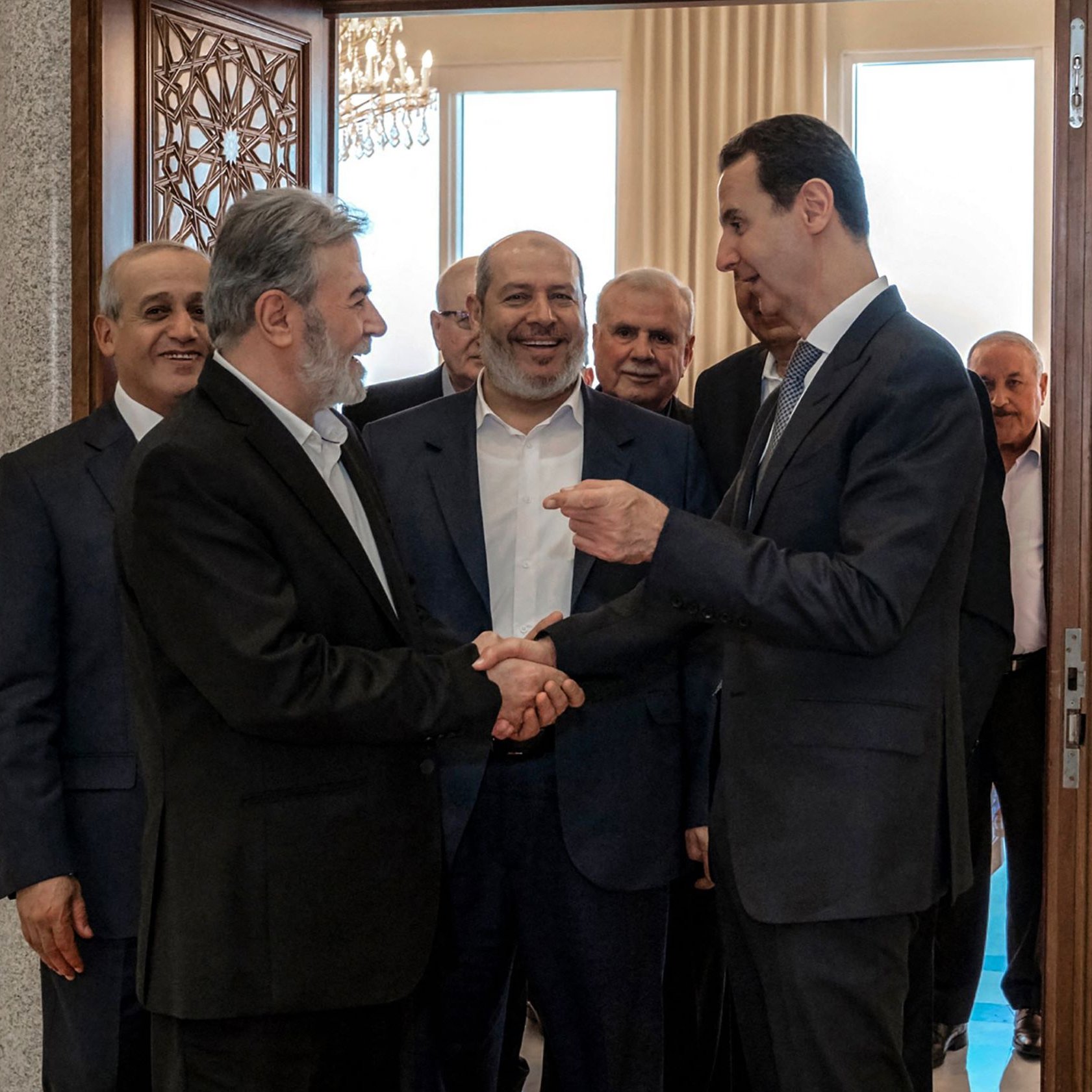 در این تصویر، چند مرد در یک اتاق ملاقات کرده‌اند. در جلو، دو نفر رو به روی هم ایستاده‌اند و با هم دست می‌دهند. فردی که سمت راست است ظاهراً در حال صحبت کردن و لبخند زدن است. دیگر افراد در پس‌زمینه نیز به این دو نفر نگاه می‌کنند و به نظر می‌رسد که در حال گفتگو یا مکالمه‌ای دوستانه هستند. فضا دارای نور طبیعی خوبی است و طراحی داخلی اتاق مدرن و شیک به نظر می‌رسد.