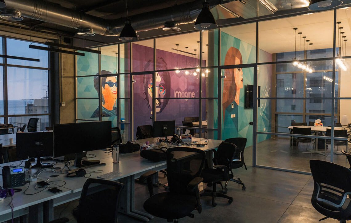 L'image montre un espace de bureau moderne avec de grandes fenêtres offrant une vue sur l'extérieur. Les murs sont décorés de peintures murales colorées représentant des visages stylisés. Le mobilier est minimaliste et fonctionnel, avec des tables de travail et des chaises ergonomiques. L'éclairage est tamisé, créant une ambiance chaleureuse malgré le design contemporain. On peut également voir des ordinateurs sur les bureaux, indiquant que l'espace est utilisé pour le travail.