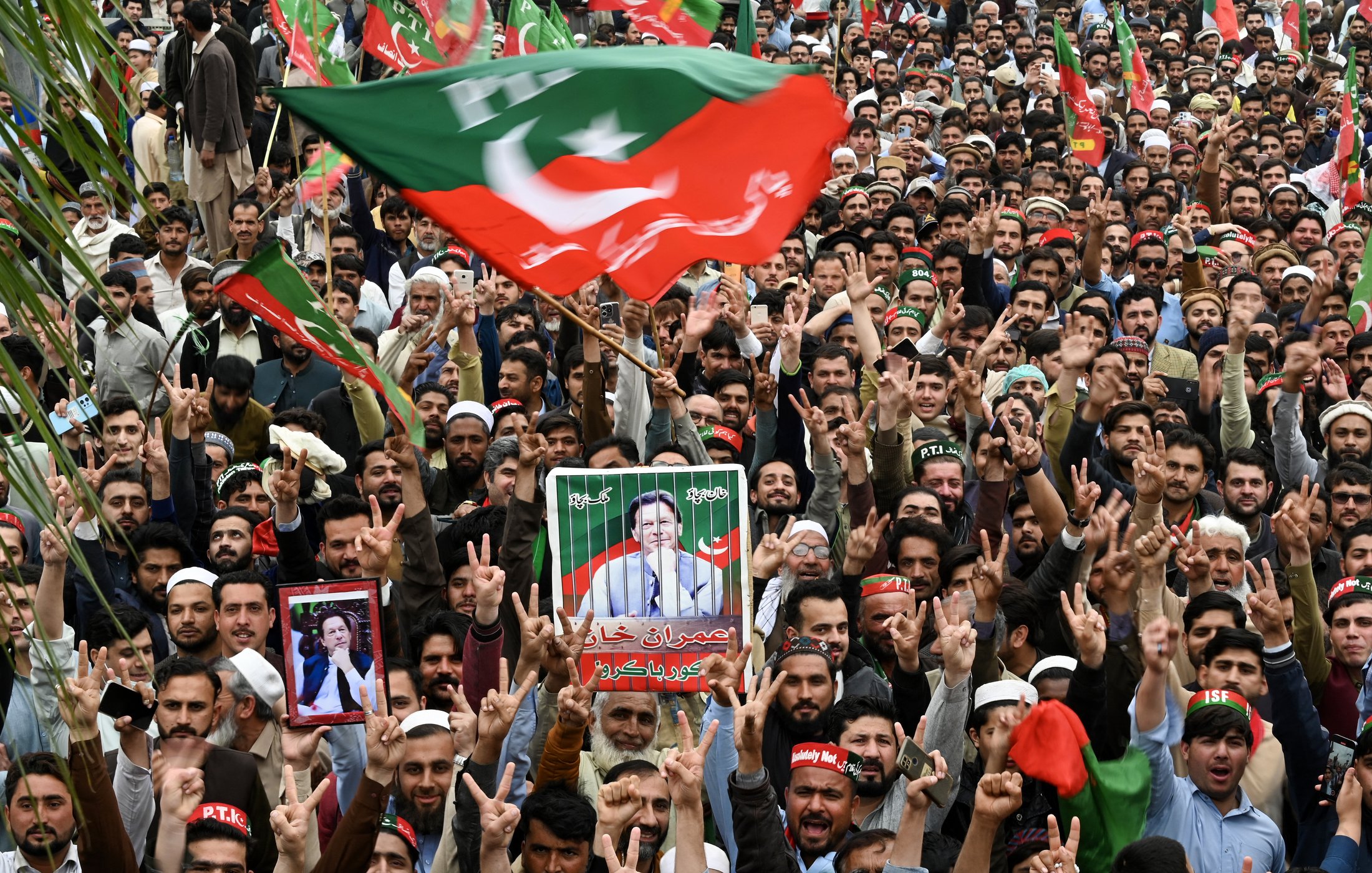L'image montre une grande foule rassemblée lors d'un rassemblement. De nombreuses personnes brandissent des drapeaux, en particulier le drapeau du Parti Pakistan Tehreek-e-Insaf (PTI). On peut voir des manifestants levant les bras avec des gestes de victoire, ainsi que des pancartes et des portraits de leaders politiques. L'ambiance semble dynamique et engageante, avec des cris et des slogans de la part des partisans.