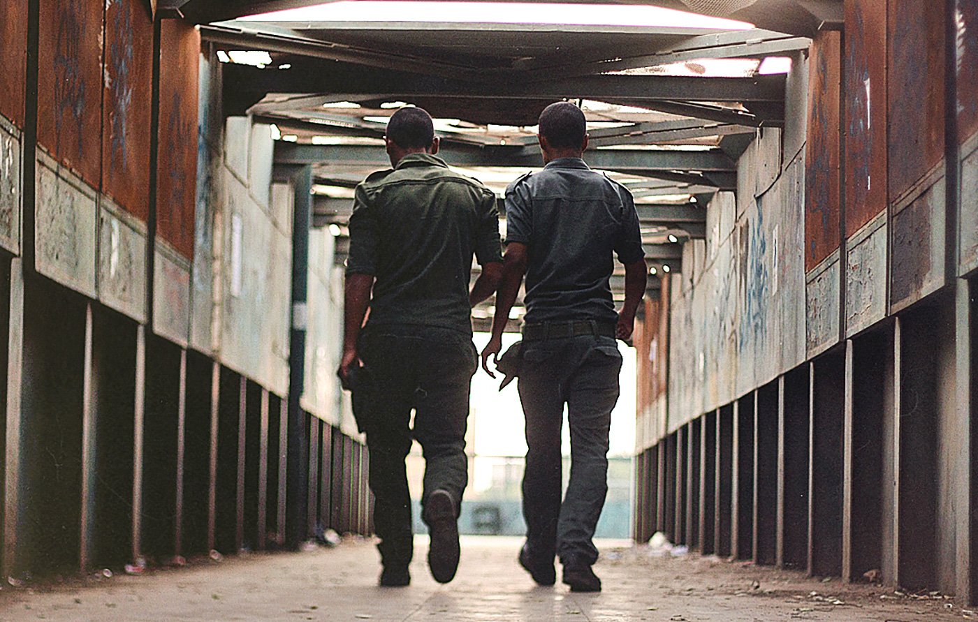 L'image montre deux hommes marchant côte à côte dans un passage couvert. Ils portent des uniformes de couleur sombre et se déplacent vers la sortie, avec leurs silhouettes se détachant contre la lumière. Les murs du passage semblent usés, avec des graffitis visibles, et le sol est pavé. L'atmosphère est à la fois urbaine et légèrement mélancolique, donnant une impression d'intimité entre les deux personnages.