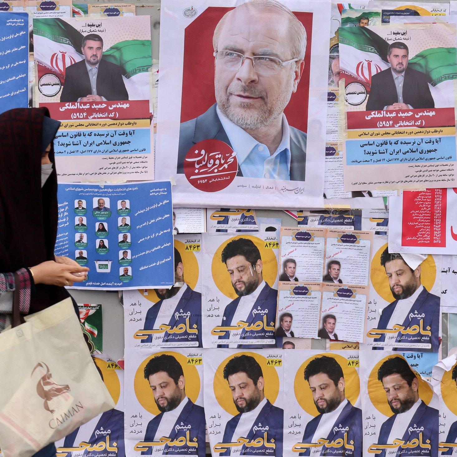 L'image montre un mur recouvert de diverses affiches électorales. On peut voir plusieurs portraits de candidats, accompagnés de slogans et d'informations. Au premier plan, une femme portant un hijab et tenant un sac semble marcher devant ces affiches, ce qui suggère une ambiance électorale. Les couleurs des affiches sont vives et variées, ajoutant à l'aspect animé de la scène.