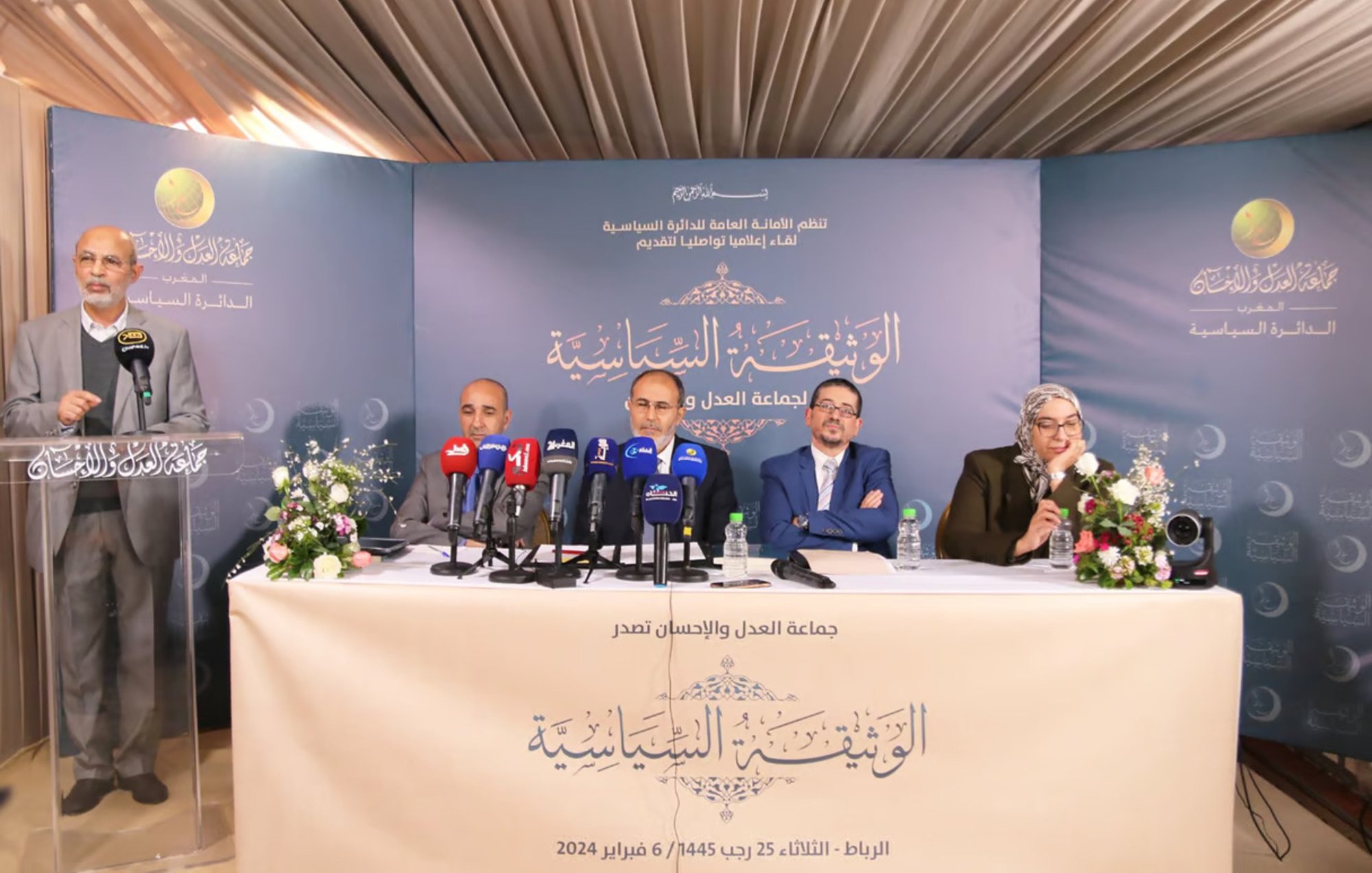 La imagen muestra a un grupo de cuatro personas sentadas en una mesa de prensa durante un evento. Hay micrófonos de diferentes medios de comunicación frente a ellos, y el ambiente parece ser formal. La persona de pie, a la izquierda, parece estar dando un discurso o presentación. El fondo incluye un cartel con texto en árabe, y hay arreglos de flores sobre la mesa. La escena sugiere que se está llevando a cabo una conferencia o reunión importante.