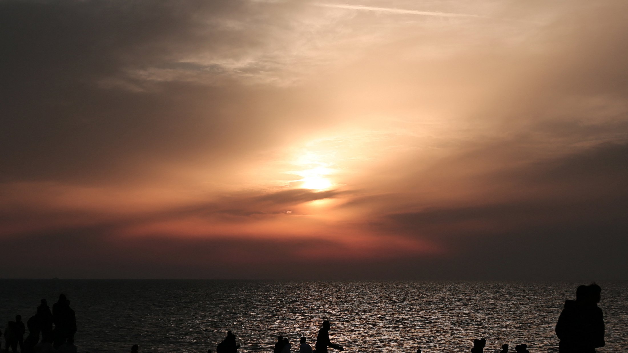 L'immagine mostra una scena al tramonto sulla spiaggia. Il cielo è dipinto di tonalità calde, con sfumature di arancione e rosa, mentre il sole si avvicina all'orizzonte. In primo piano, si vedono diverse silhouette di persone che camminano lungo la riva, alcune sedute o accoccolate sulla sabbia. La superficie dell'acqua riflette i colori del cielo, creando un'atmosfera calma e suggestiva.