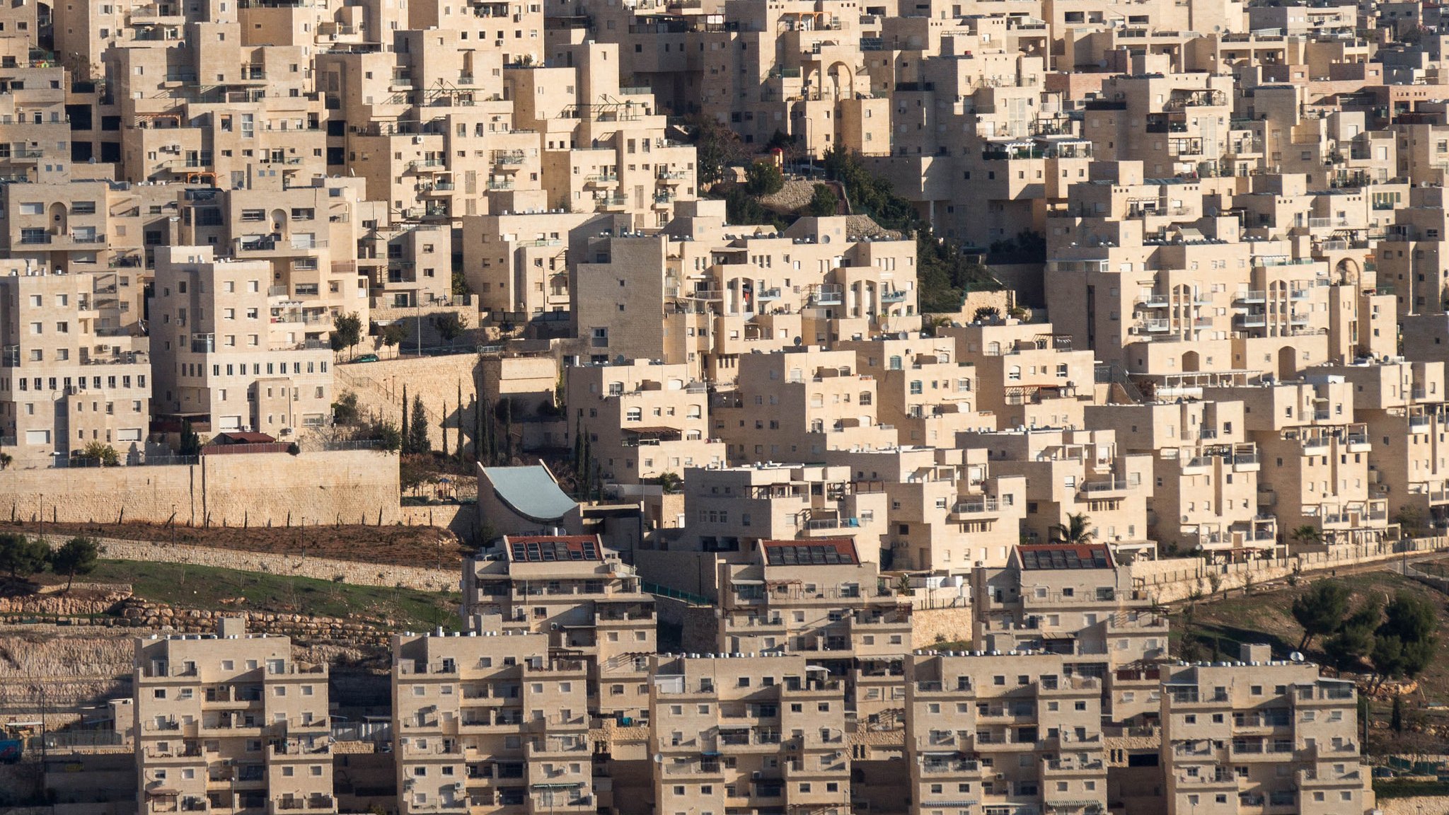 L'image présente un paysage urbain dense, constitué de nombreux bâtiments aux formes géométriques. Les structures sont principalement de couleur claire, avec des nuances de beige et de blanc. On peut observer des immeubles empilés les uns sur les autres, créant une apparence presque uniforme. Le terrain autour des bâtiments semble légèrement vallonné, avec des zones de végétation sporadique. Il y a également des éléments architecturaux distincts, tels qu'un toit qui se distingue parmi l'ensemble des constructions. L'ensemble donne une impression de modernité et d'urbanisation.