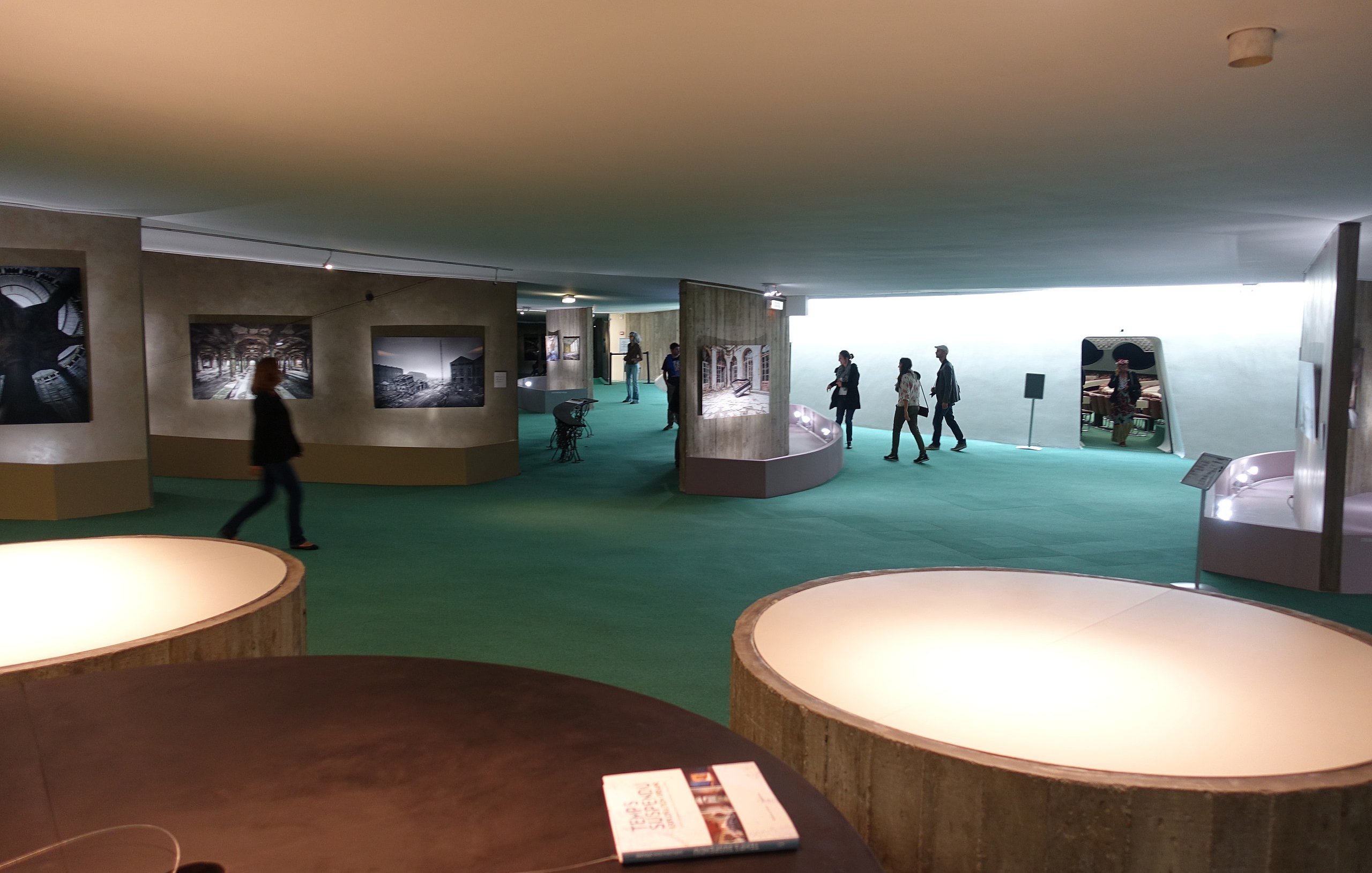 L'image présente un espace d'exposition, probablement dans un musée. On peut y voir des murs de couleur claire avec des photographies accrochées. Le sol est recouvert d'un tapis vert, créant une atmosphère accueillante. Des visiteurs se déplacent dans cet espace, explorant les œuvres exposées. Au premier plan, il y a plusieurs tables ou surfaces circulaires, apparemment utilisées pour des brochures ou d'autres informations. L'éclairage est doux et met en valeur les œuvres sur les murs.