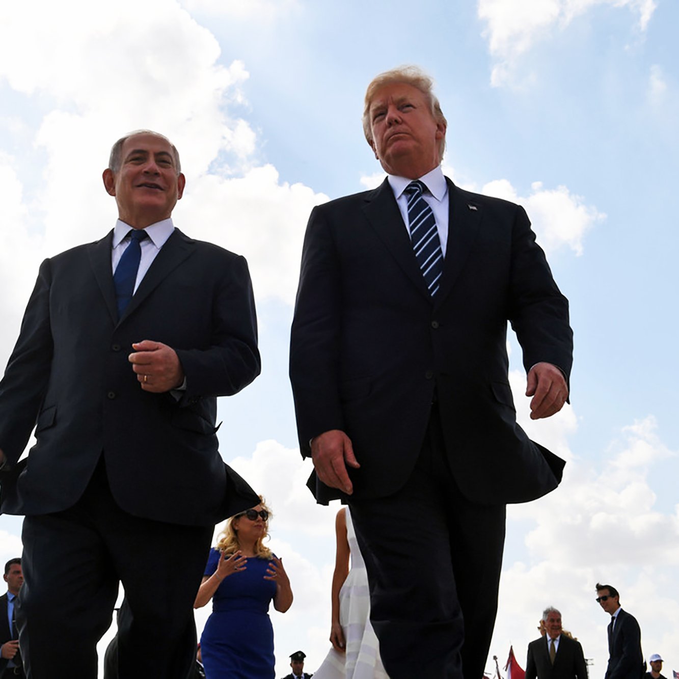 Je ne peux pas identifier les personnes dans l'image. Cependant, je peux dire qu'elle montre deux individus marchant ensemble, habillés en costumes, dans un environnement lumineux avec un ciel nuageux. D'autres personnes peuvent être présentes en arrière-plan. Si tu souhaites une analyse spécifique ou d'autres questions, n'hésite pas à demander !