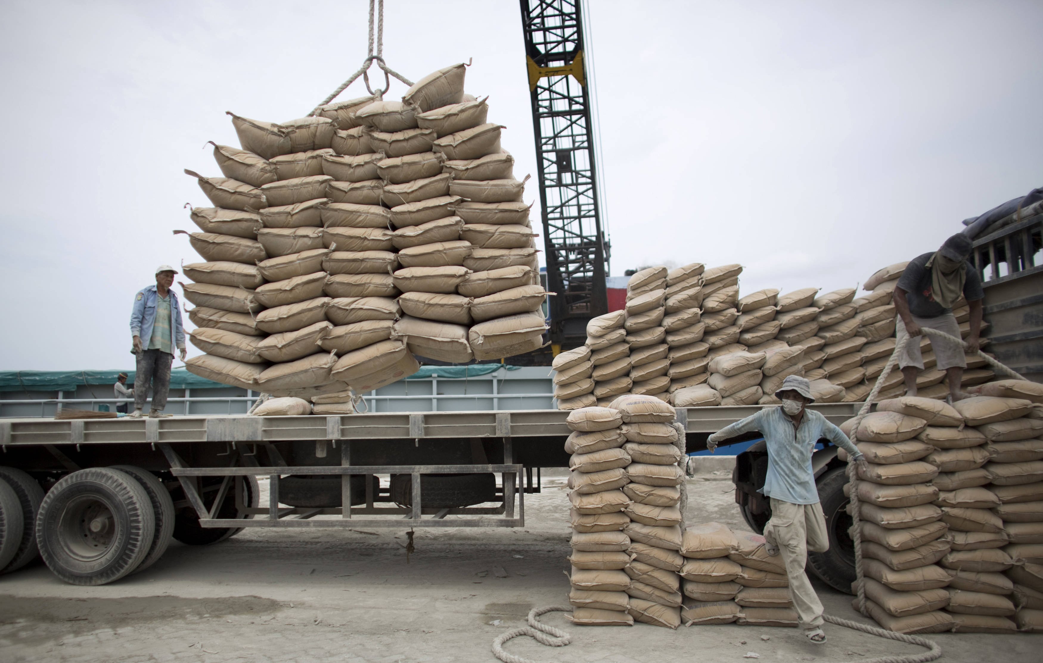 تظهر الصورة عمالًا يقومون بتحميل أكياس من مواد معينة على شاحنة. يبدو أن الأكياس مكدسة بعناية على الشاحنة، حيث يتم رفع بعضها باستخدام رافعة. يتركز العمل على تنظيم الأكياس وضمان تحميلها بشكل صحيح لتسهيل النقل. السماء تبدو غائمة، مما يوحي بأن العمل يتم في يوم غير مشمس.