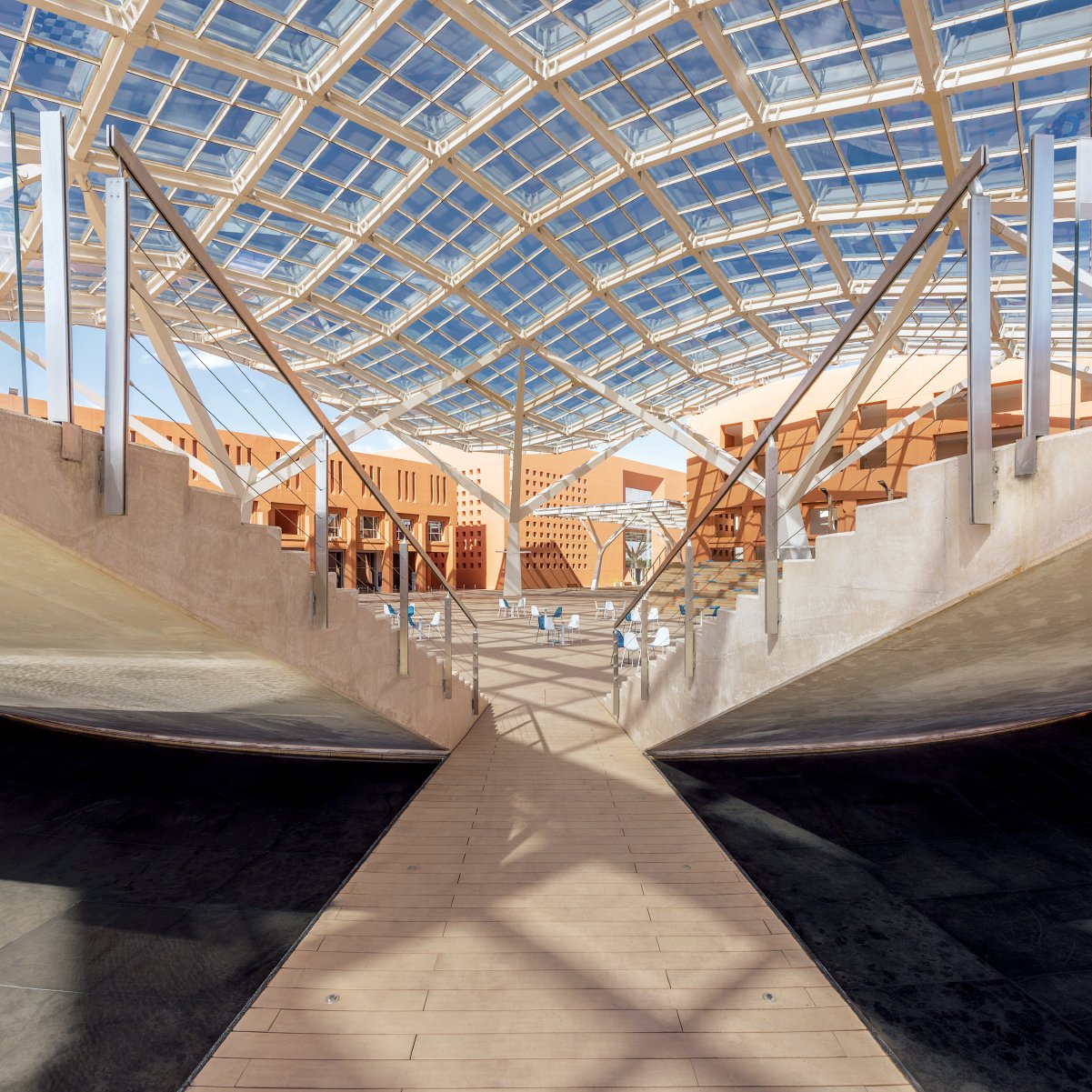 L'immagine mostra un'architettura moderna caratterizzata da una struttura ampia e luminosa. La copertura in vetro permette alla luce naturale di filtrare, creando un ambiente aperto e arioso. Le pareti laterali presentano un design minimalista, con elementi di legno e cemento che si combinano armoniosamente. Al centro, una passerella in legno collega le diverse aree, accentuando una sensazione di continuità e apertura nello spazio. La scena è caratterizzata da linee pulite e forme geometriche, suggerendo un design innovativo e funzionale.