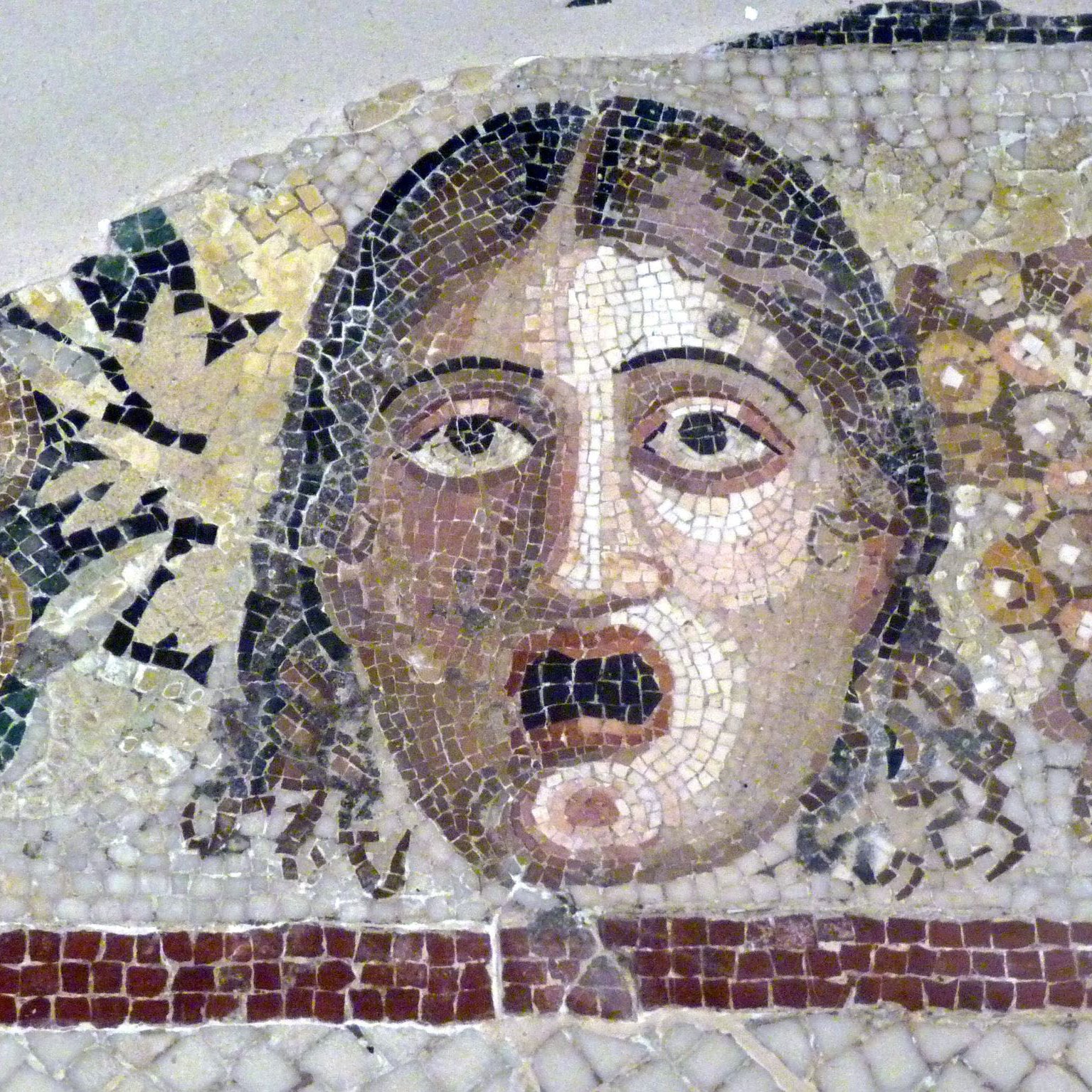 L'image présente un ancien mosaïque représentant un visage humain. Le caractère exprime une émotion marquée, semblant étonné ou effrayé. Autour du visage, il y a des éléments décoratifs, notamment des grappes de raisin et des fruits, qui ajoutent une touche colorée et contrastent avec le fond neutre de la mosaïque. La technique utilisée évoque la richesse de l'art ancien et son attention aux détails.