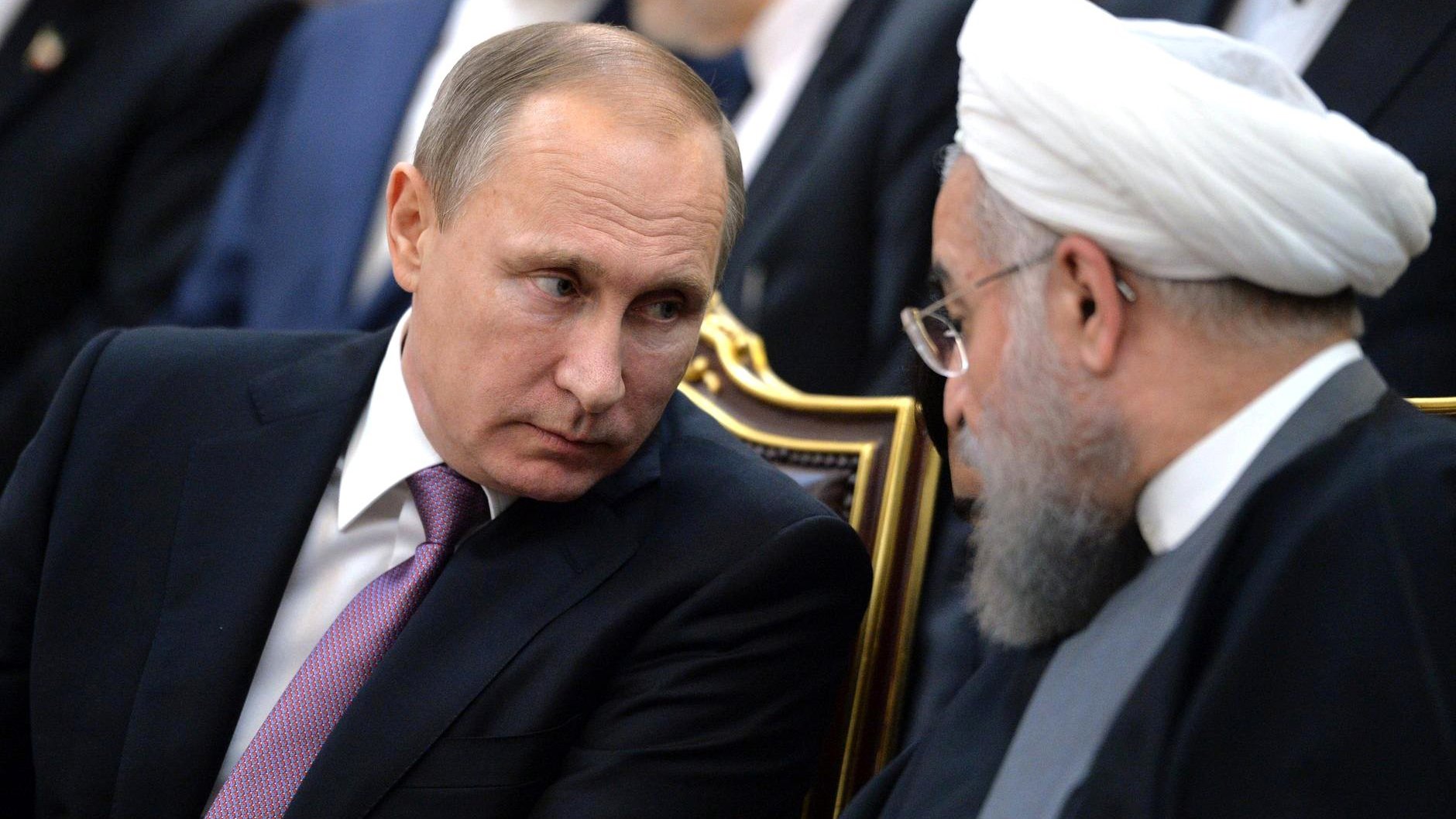 The image depicts two men engaged in a conversation. One is seated with a serious expression, wearing a dark suit and a light-colored tie. The other man, also seated, has a beard and wears traditional clerical attire, including a white turban. They appear to be in a formal setting, possibly during a meeting or event, with other individuals visible in the background. The focus is on their interaction and expressions.