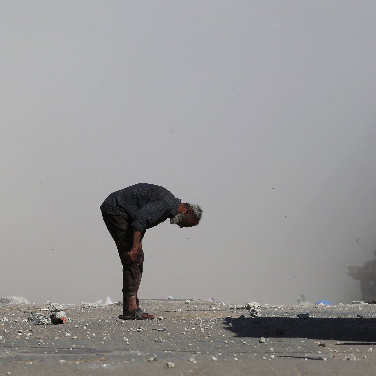 مردی مسن با کمر خمیده در مکانی خاکی و غبارآلود ایستاده است. اطرافش صحنه‌ای ویران وجود دارد.