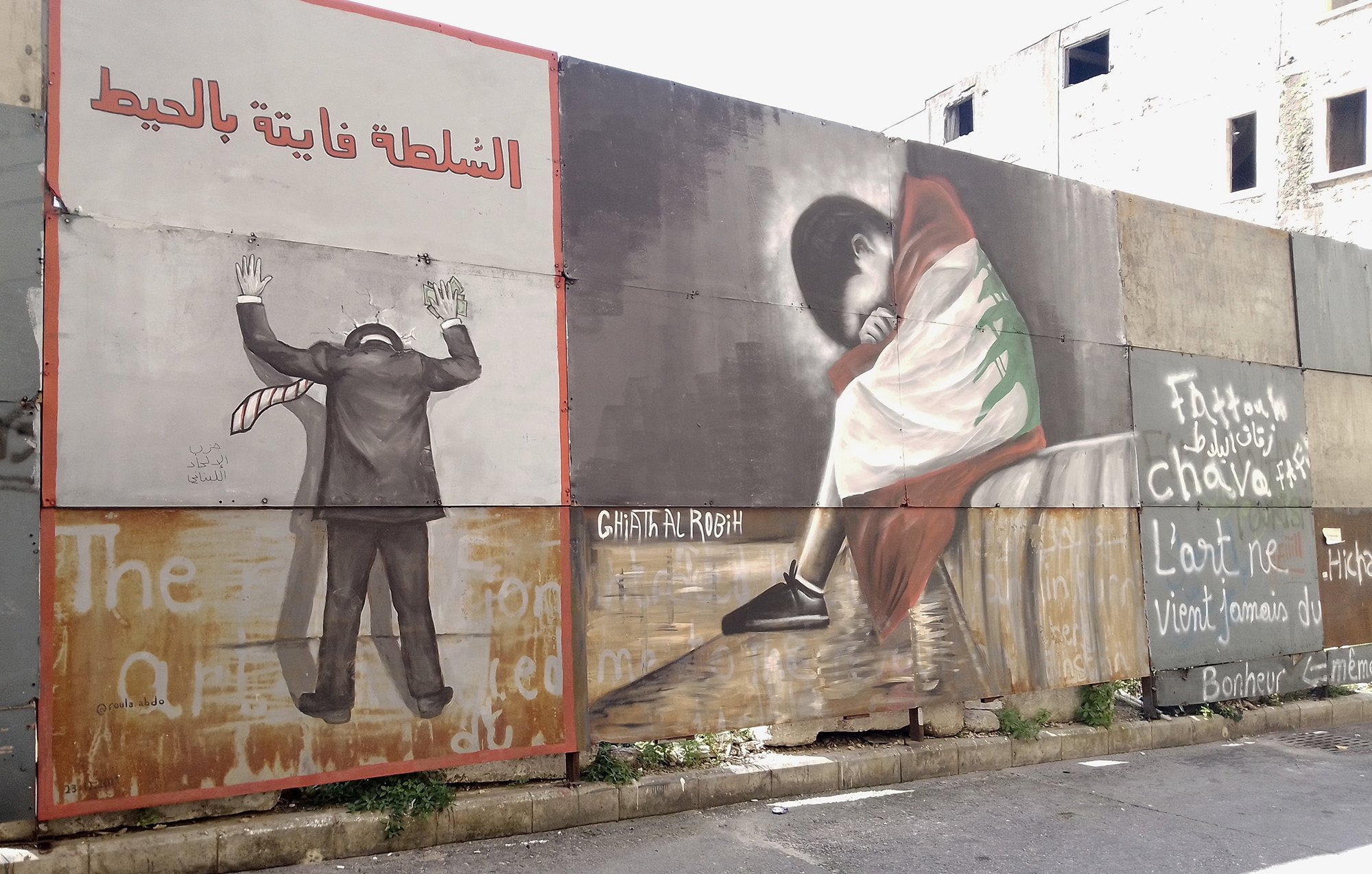 L'image montre un mur recouvert de plusieurs peintures murales. À gauche, on voit une figure humaine en costume, les mains levées, comme en signe de désespoir ou de soumission, avec une inscription en arabe qui pourrait évoquer des thèmes de répression. À droite, une autre figure, probablement d'une personne en train de pleurer ou de s'asseoir tristement, est représentée, vêtue d'une robe aux couleurs d'un drapeau, ce qui pourrait symboliser une forte connexion à une identité culturelle ou nationale. Le fond comporte également des mots ou des phrases en français et en arabe, ajoutant une dimension de commentaire social ou politique à l'œuvre. L'ensemble évoque des thèmes de lutte, d'identité et d'émotion.