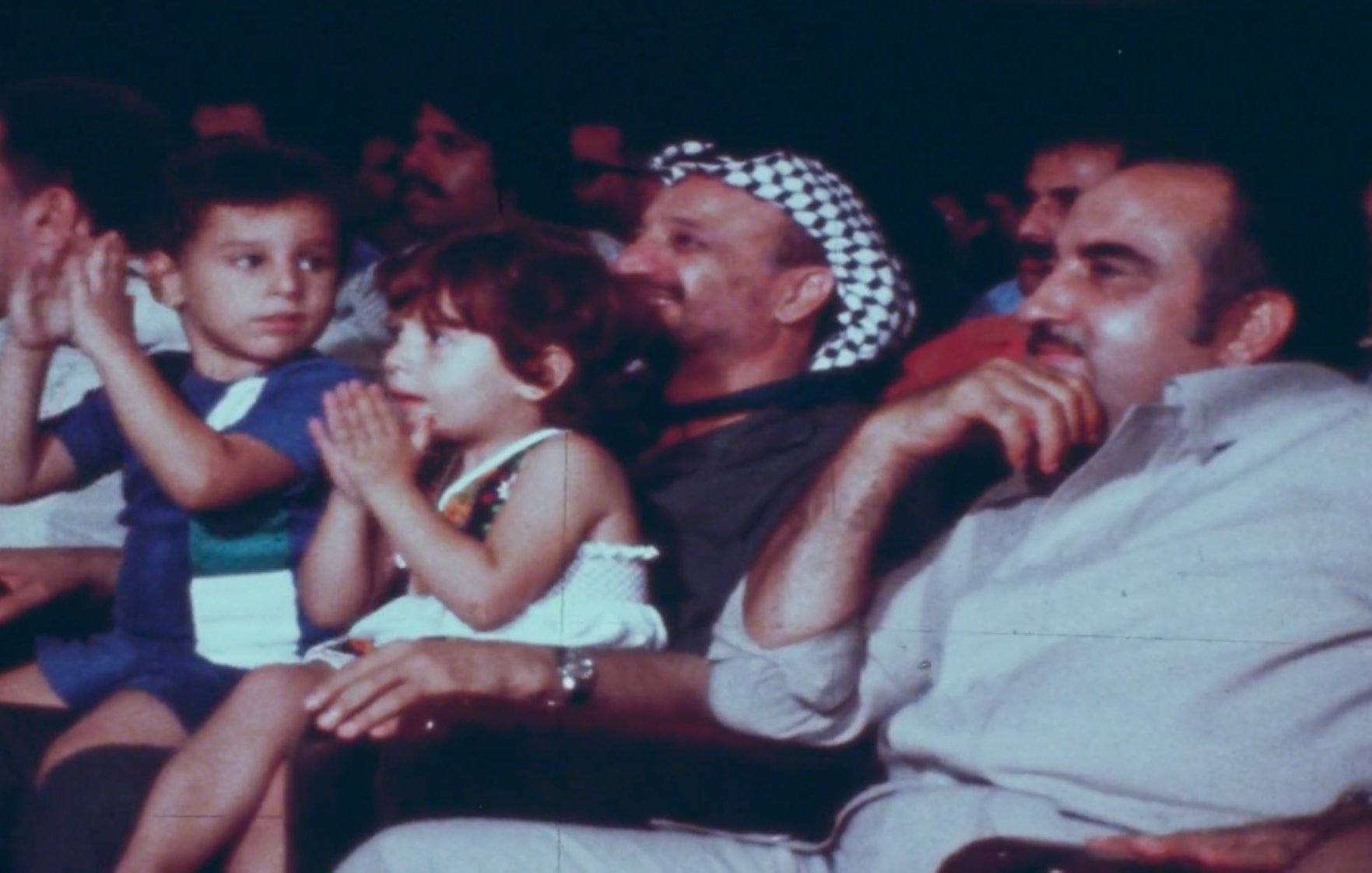 L'image montre un groupe de personnes assises dans une salle, probablement lors d'un événement. Au premier plan, deux enfants, un garçon et une fille, semblent applaudir. Le garçon porte un maillot de bain et la fille est habillée avec une petite robe. Derrière eux, un homme portant un keffieh (le couvre-chef traditionnel) sourit. À sa droite, un autre homme est assis, regardant également vers le devant. L'ambiance paraît festive et conviviale.