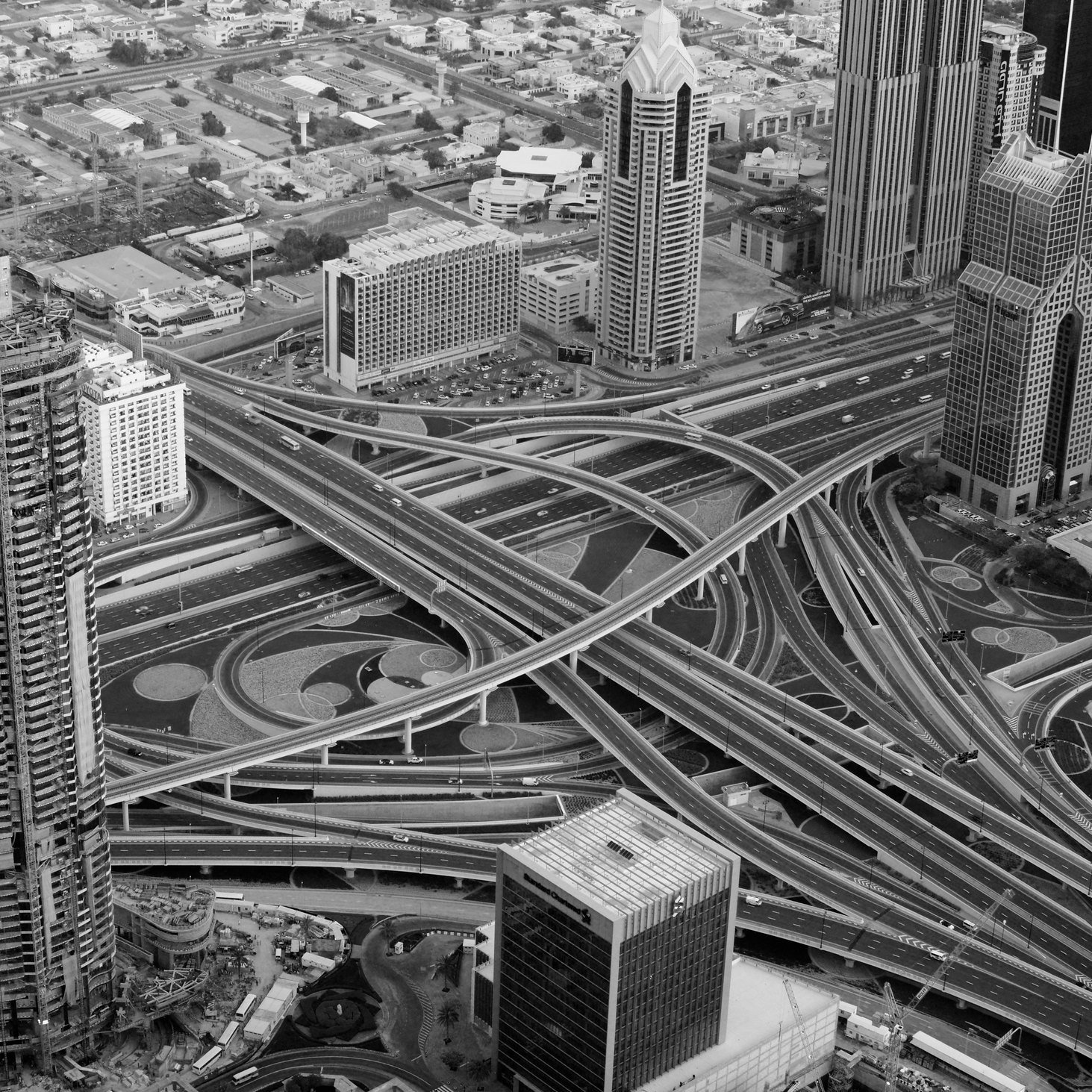 L'image présente une vue aérienne d'une métropole moderne, probablement Dubaï. On y voit de grandes tours élégantes et contemporaines, ainsi qu'un réseau complexe de routes et d'autoroutes qui s'entrelacent. Le contraste en noir et blanc accentue les formes géométriques des bâtiments et des routes, créant une impression de dynamisme et d'organisation. Des zones urbaines et dégagées suggèrent une planification urbaine sophistiquée.