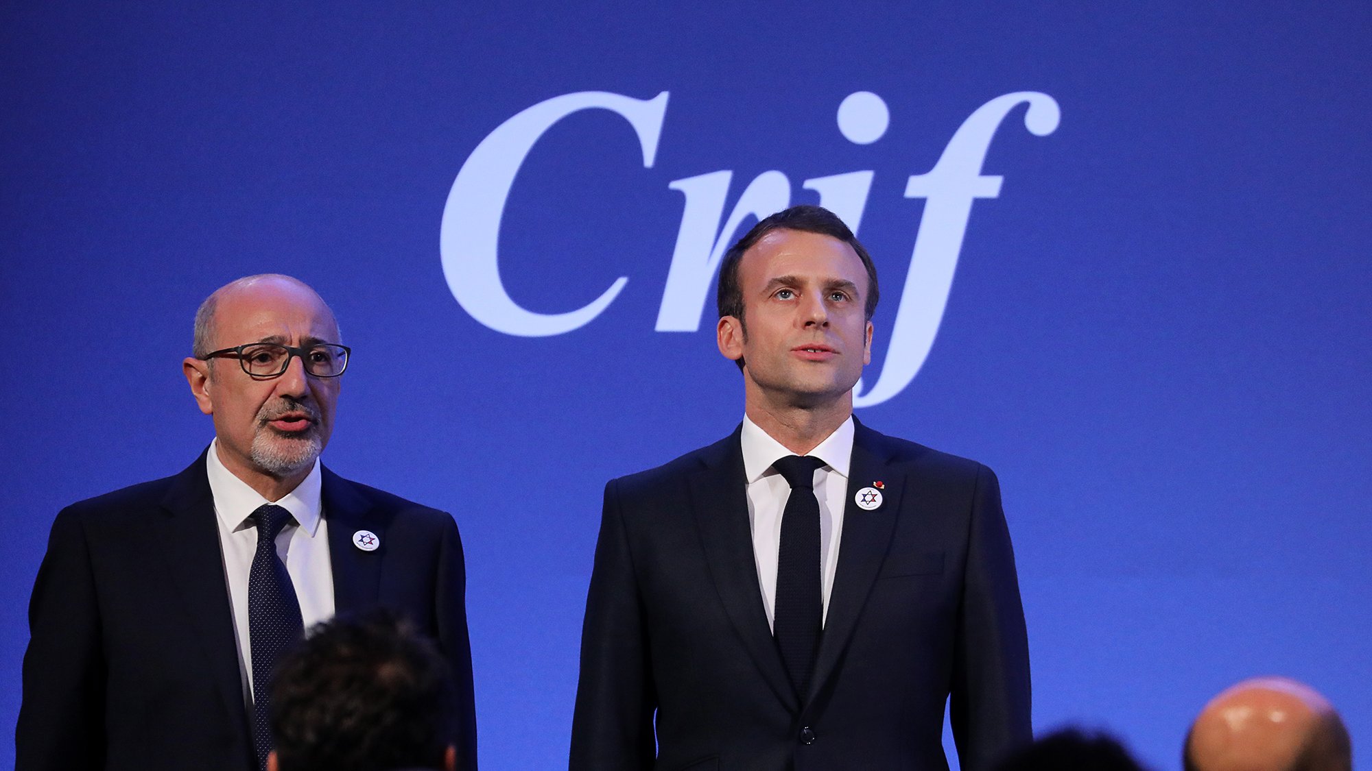 L'image montre deux personnes sur scène, probablement lors d'un événement officiel. À gauche, un homme porte des lunettes et un costume sombre, tandis qu'à droite, un homme, probablement un dirigeant, se tient debout en costume noir avec une cravate, affichant une expression sérieuse. En arrière-plan, le mot "Crif" est visible, ce qui suggère que l'événement est lié à cette organisation. L'atmosphère semble solennelle et professionnelle.