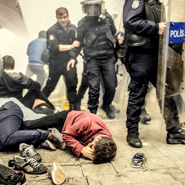 L'image montre une scène de manifestation ou d'émeute. On peut voir plusieurs personnes au sol, certaines semblant blessées ou cernées par des forces de police. Des policiers en tenue anti-émeute avancent dans une atmosphère de chaos, avec des gaz lacrymogènes obscurcissant l'air. L'environnement paraît tendu, témoignant d'un conflit entre manifestants et autorités.