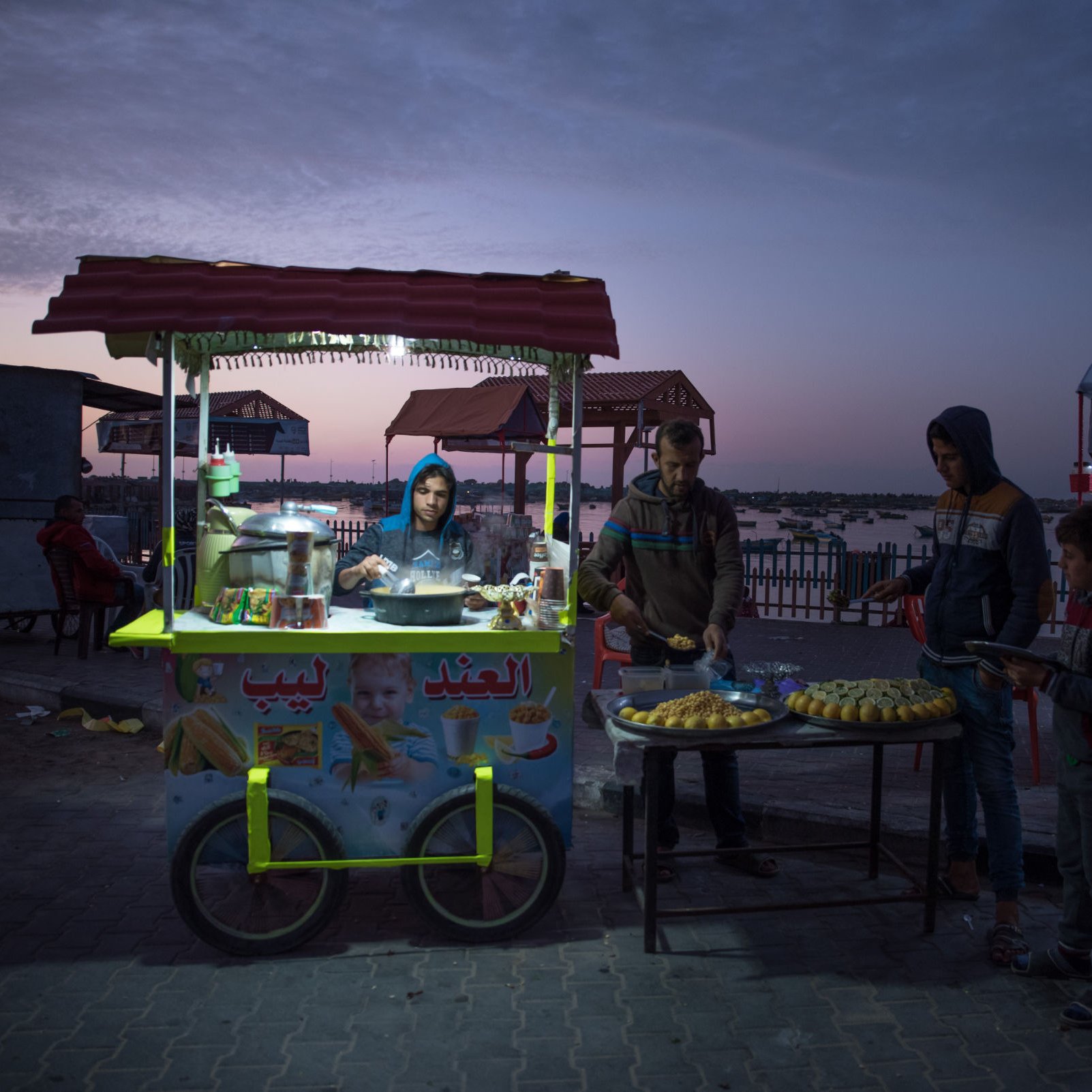 L'image montre un stand de nourriture de rue à la tombée de la nuit, avec un ciel coloré par le crépuscule. Au premier plan, un vendeur prépare des aliments, tandis que plusieurs jeunes clients attendent. Le stand est décoré avec des affiches et contient des produits alimentaires, tels que des pâtisseries. En arrière-plan, on peut apercevoir d'autres stands illuminés et des chaises, créant une ambiance conviviale.