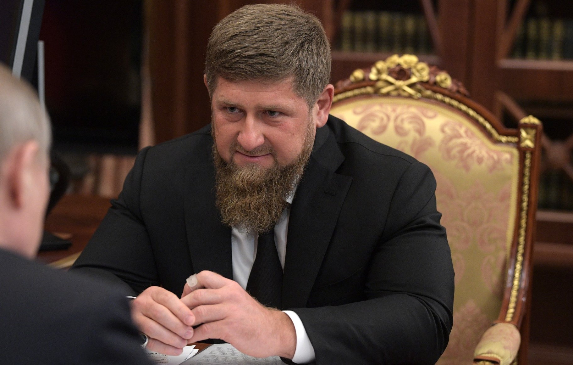 الصورة تظهر رجلًا يجلس على طاولة، وهو يرتدي بدلة رسمية سوداء. يتمتع بلحية كثيفة ويمسك بيديه شيئًا ما. خلفه تظهر تفاصيل من مكتب فاخرة، مثل الأثاث المزخرف والمكتبات. يبدو أنه في اجتماع أو مناقشة مع شخص آخر.