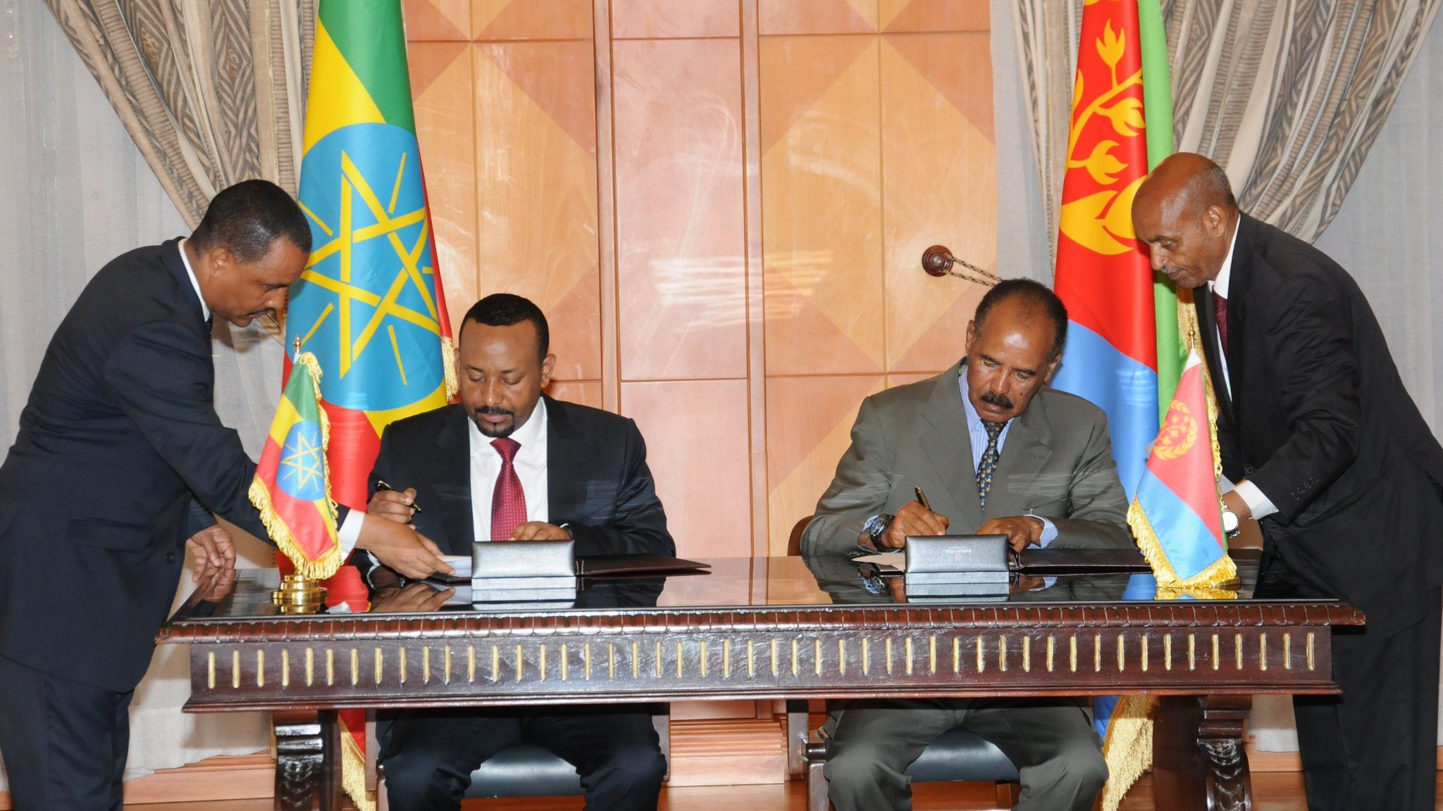 تظهر الصورة لحظة توقيع اتفاقية أو بروتوكول بين ممثلين من دولتي إثيوبيا وإريتريا. يجتمع أربعة رجال حول طاولة، حيث يقوم اثنان منهم بالتوقيع على مستندات برفقة الأعلام الوطنية للدولتين. خلفهم توجد ستائر وأثاث رسمي يضفي أجواء من الرسمية على المناسبة. يعبّر المشهد عن تعاون أو اتفاقية مهمة بين البلدين.
