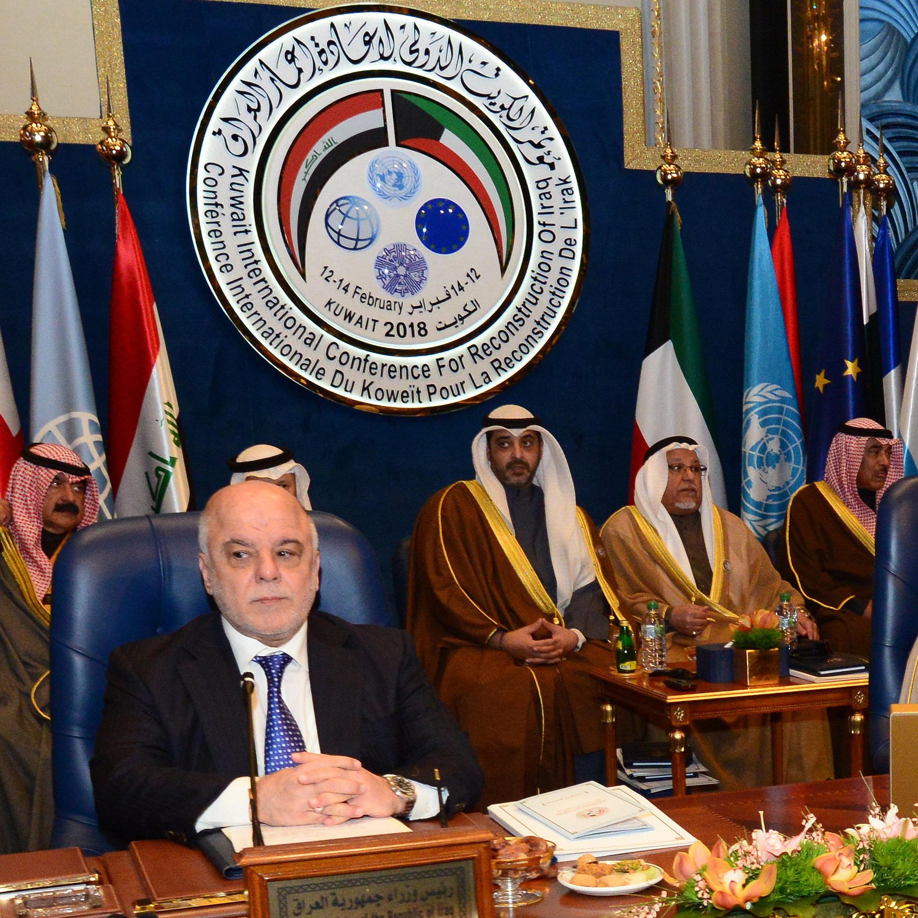 The image depicts a formal conference setting, featuring several dignitaries sitting in a room with flags from various nations displayed prominently in the background. The attendees are dressed in traditional attire, indicating a Middle Eastern cultural context. A large emblem or logo is visible above them, likely representing the conference's purpose, which involves international cooperation or reconstruction efforts. Among the seated figures, one appears to be a prominent leader or official, positioned at the forefront of the image. The overall atmosphere suggests a significant meeting aimed at addressing important regional or global issues.
