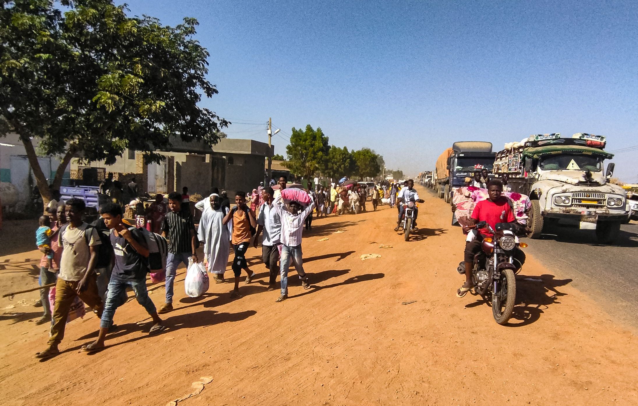 L'image montre une scène de rue animée dans une zone urbaine. Sur le côté gauche, on voit un arbre et des bâtiments, tandis que des gens marchent le long de la route en terre. Parmi eux, certains portent des sacs ou des charges sur la tête. Des véhicules, y compris des camions et des motos, circulent sur la route, créant une ambiance dynamique. L'éclairage semble être en plein jour, suggérant une activité quotidienne typique dans cette communauté.