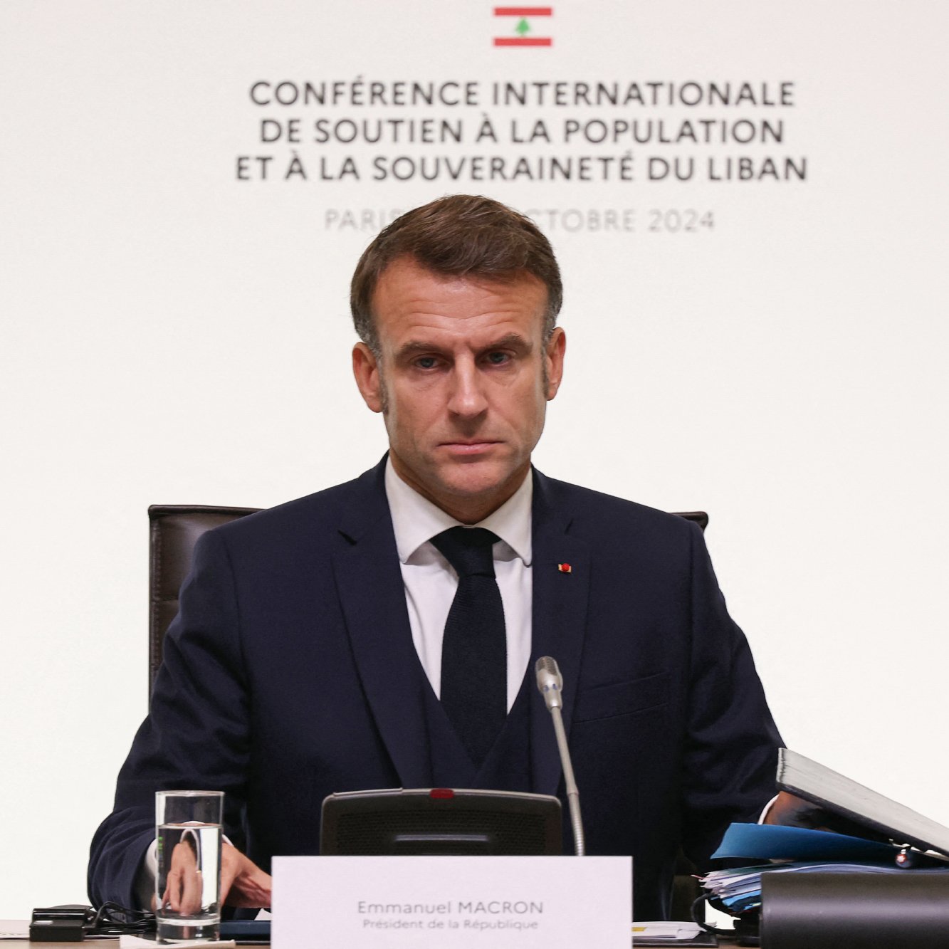 L'image montre un homme assis à une table lors d'une conférence. Il est habillé en costume sombre et porte une cravate. L'arrière-plan présente des inscriptions en français, indiquant qu'il s'agit d'une conférence internationale soutenant la population et la souveraineté du Liban, à Paris en octobre 2024. L'homme semble concentré et sérieux, regardant vers l'avant. Sur la table, on peut voir un verre d'eau et des documents.