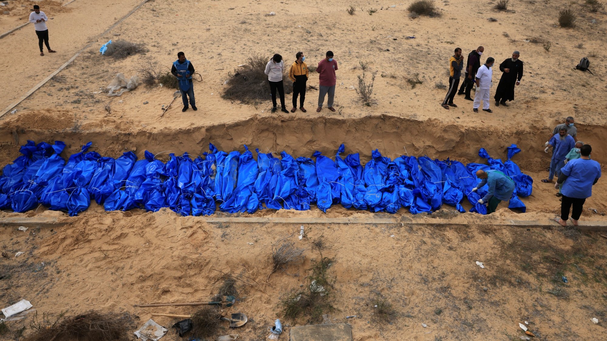 Des corps enveloppés de bleu sont placés dans une fosse, entourés de personnes en deuil.