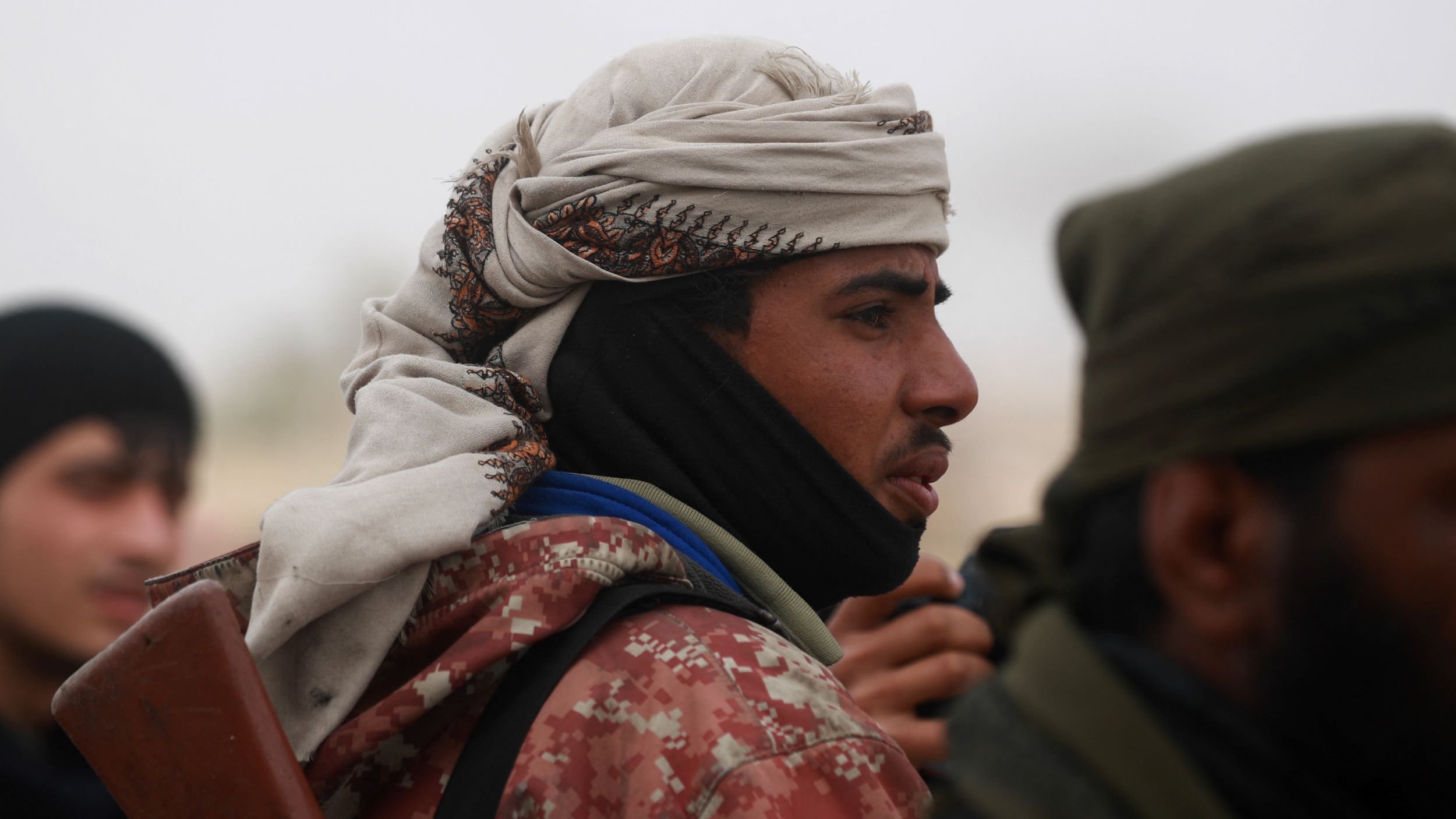 در این تصویر، یک فرد در حالتی نزدیک به دوربین قرار دارد. او کلاهی بر سر دارد و چهره‌اش جزئیاتی را نشان می‌دهد که به او ظاهری خاص می‌دهد. لباسش نیز شامل یک روپوش است که الگوهای خاصی دارد. در پس‌زمینه، افرادی دیگر نیز دیده می‌شوند. جو تصویر احتمالاً جدی و نظامی است.