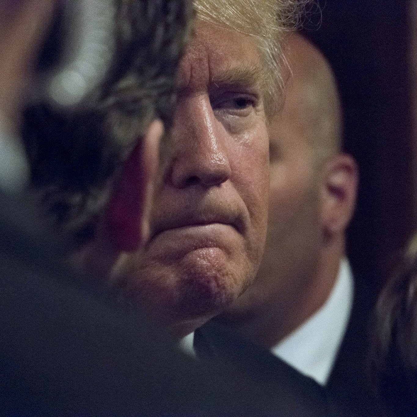 در این تصویر، یک مرد با موهای روشن و کمی حالت خمیده سر دیده می‌شود. او در حالتی contemplative به نظر می‌رسد و گونه‌ها و خطوط صورتش مشخص است. در اطراف او چندین فرد دیگر مشاهده می‌شوند که به نظر می‌رسد در حال گفتگو یا مراقبت از او هستند. پس‌زمینه تصویر تاریک و مبهم است، که تمرکز را بر روی چهره او بیشتر می‌کند. این تصویر احساس جدیت و تفکر عمیق را منتقل می‌کند.