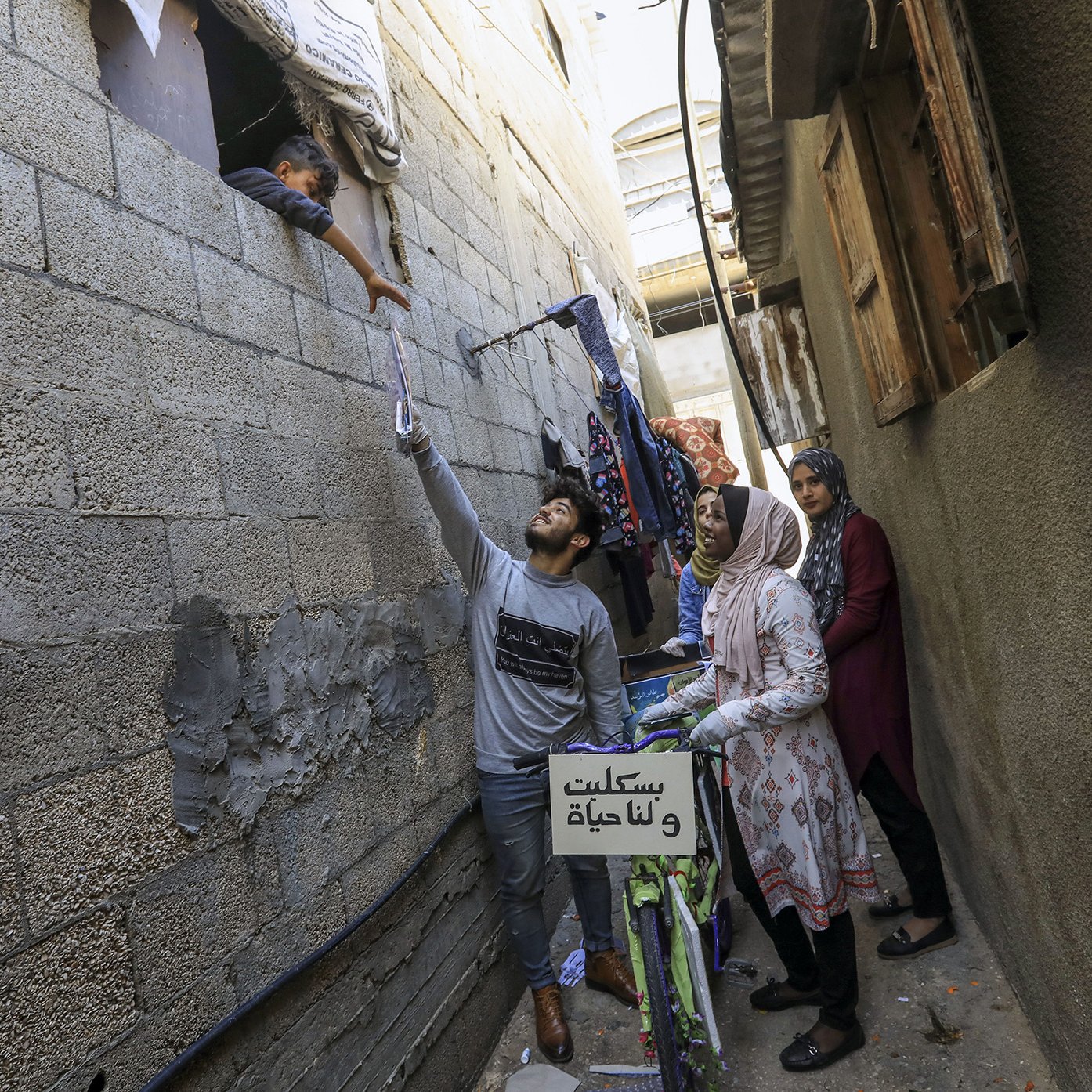 L'image montre une scène dans une ruelle étroite, probablement dans un quartier urbain. On y voit un jeune homme qui tend la main vers une fenêtre, où un enfant semble interagir avec lui. À l'arrière-plan, une femme, probablement la mère de l'enfant, observe la scène. Une autre femme se tient à côté, regardant également. Des vêtements sont suspendus à l'extérieur, ajoutant une touche de vie quotidienne à l'image. Une bicyclette est visible, avec un panneau sur le guidon. L'atmosphère semble chaleureuse et familiale, malgré le contexte urbain.