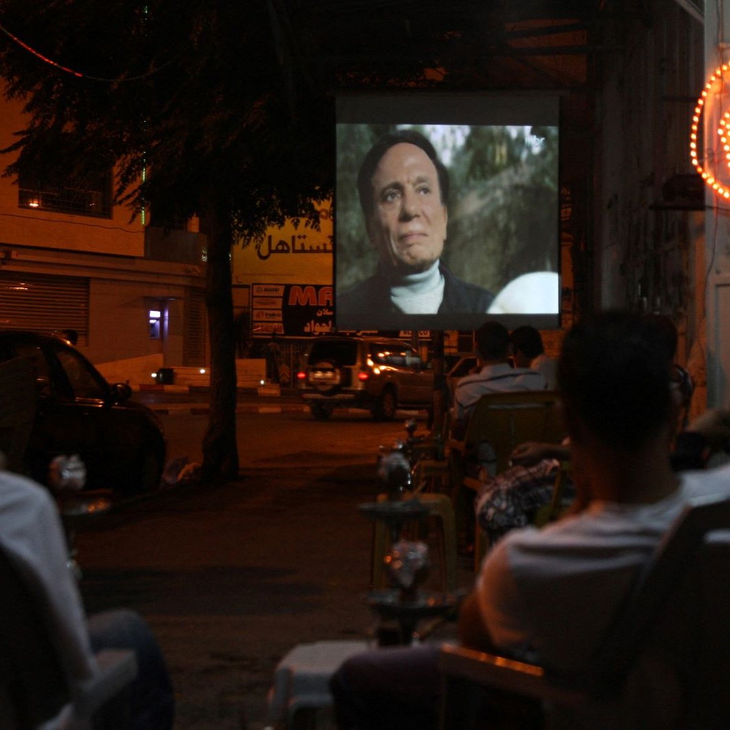 The image depicts a night scene where a group of people is seated in chairs, watching a film projected onto a large screen set up outside. The atmosphere appears relaxed, with some of the viewers enjoying drinks, possibly shisha. The area is illuminated by both the projection light and neon signage in the background, adding a lively ambiance to the setting. The scene captures a communal experience centered around outdoor movie viewing.