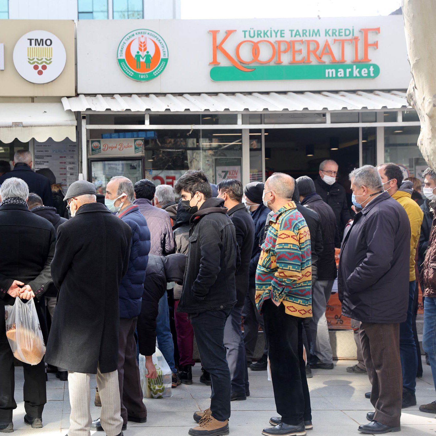 در تصویر، گروهی از مردم، عمدتاً مردان، در حال انتظار در صفی طولانی جلوی یک فروشگاه به نام "Kooperatif market" هستند. بعضی از افراد کیف و بسته‌های خرید در دست دارند و بیشتر آنها لباس‌های گرم پوشیده‌اند. در پس‌زمینه، چند ساختمان و درختان دیده می‌شود. افراد به نظر می‌رسد که منتظر خرید محصولاتی از فروشگاه هستند.