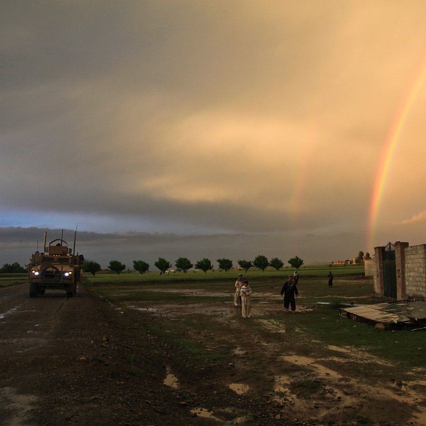 L'image montre un paysage rural sous un ciel dramatique, avec des nuages sombres et un ciel éclairci. On peut voir un arc-en-ciel double qui s'étend dans le ciel, ajoutant une touche de couleur. À gauche, un véhicule militaire est garé sur un chemin boueux, tandis qu'à droite, deux personnes marchent près d'un mur en béton. Le sol est humide, témoignant d'une récente pluie, et des arbres sont visibles en arrière-plan, soulignant une atmosphère paisible malgré la présence militaire.