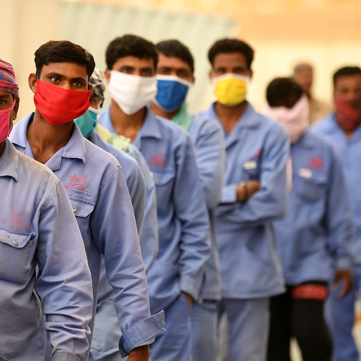 L'image montre un groupe de travailleurs masculins alignés en rang. Ils portent tous des vêtements de travail de couleur bleue et des masques de protection de différentes couleurs, notamment rouge et jaune. Les hommes semblent se préparer à entrer quelque part, et l'ambiance générale indique un contexte de travail, possiblement dans une situation où des mesures sanitaires sont en place. L'arrière-plan est flou, mettant l'accent sur les travailleurs.