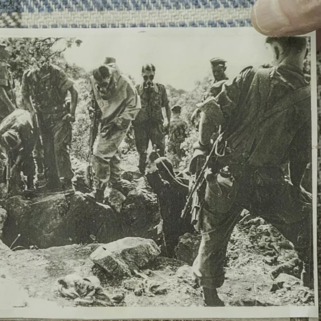L'image montre un groupe de personnes en uniforme militaire, probablement des soldats, rassemblés autour d'un trou dans le sol. Certains semblent être en train de creuser ou d'examiner le terrain, tandis que d'autres observent. L'ambiance est sérieuse, indicative d'une situation d'urgence ou de conflit. Le tout est en noir et blanc.