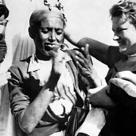 L'image montre un groupe de personnes dans une scène joyeuse et festive. Au centre, une femme souriante interagit avec un homme qui semble également être content. Ils sont entourés d'autres personnes, dont certaines affichent des expressions de bonheur. Les vêtements des personnes suggèrent une ambiance culturelle ou traditionnelle. L'atmosphère générale est celle d'une célébration ou d'un moment convivial.