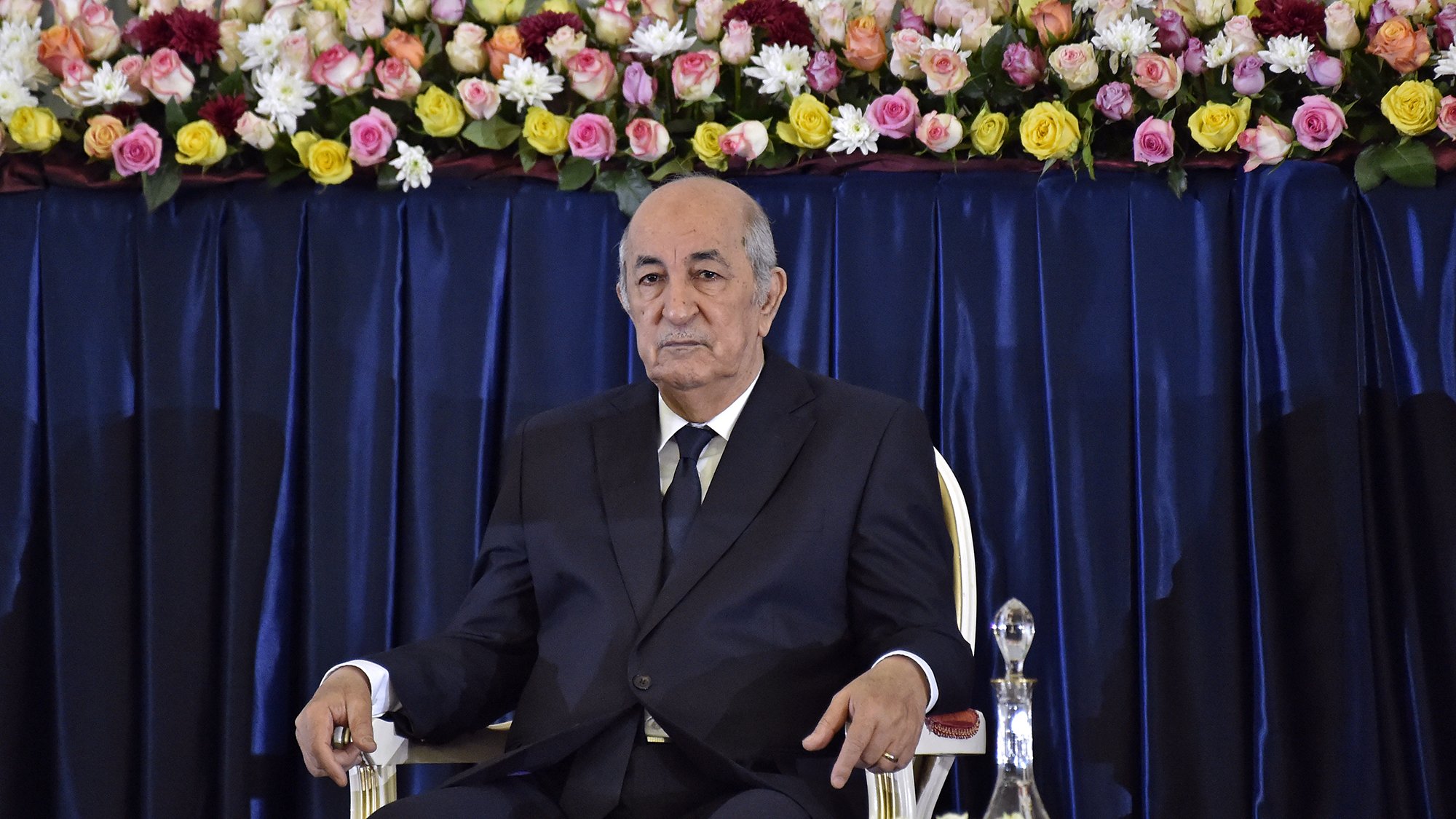 L'image montre un homme assis sur une chaise, portant un costume sombre et une cravate. Il a l'air sérieux et attentif. À l'arrière-plan, il y a une décoration florale composée de roses et d'autres fleurs qui ajoutent de la couleur à la scène. La disposition des fleurs est soignée, suggérant un cadre formel ou officiel.
