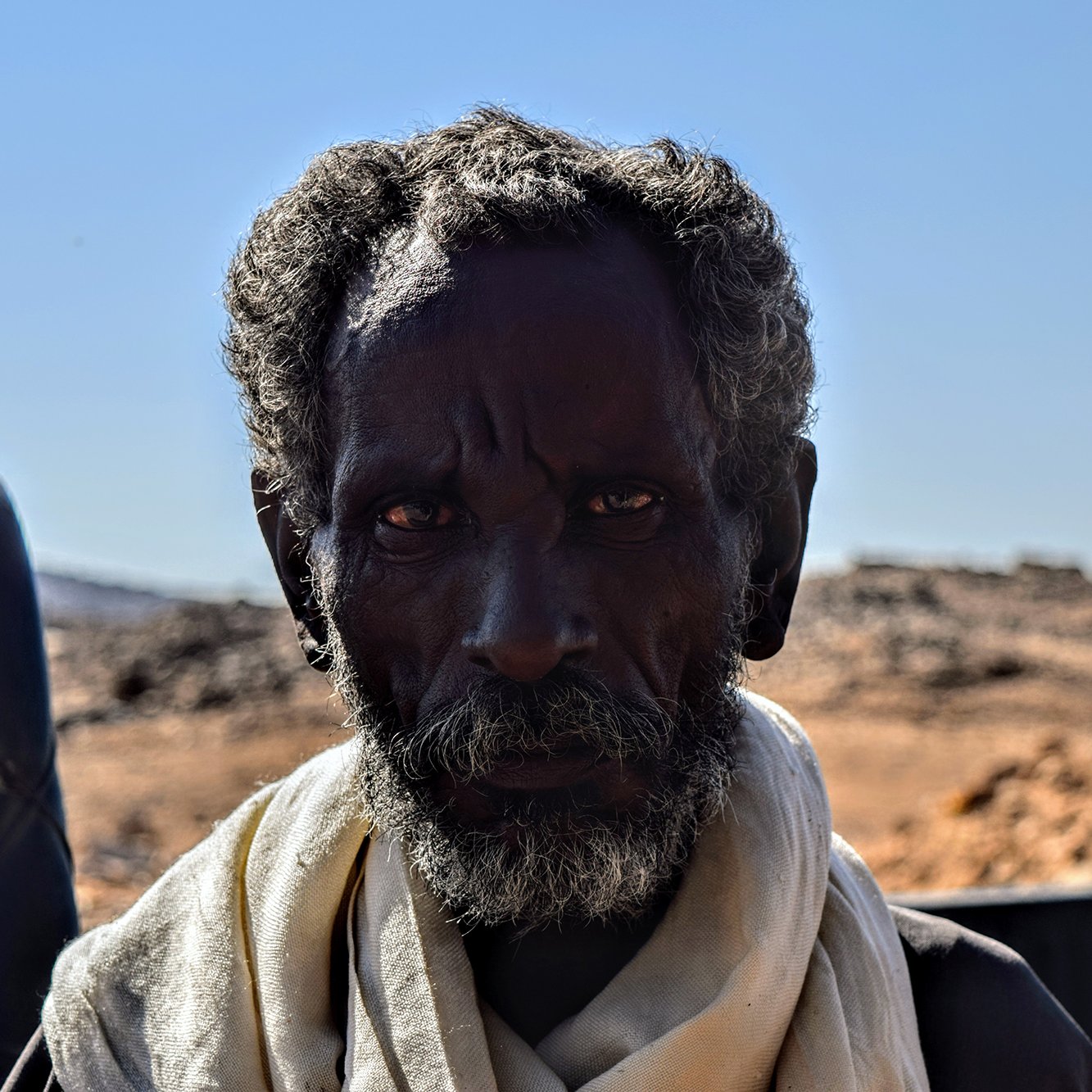 L'image montre un homme avec une peau foncée et des cheveux crépus, portant une écharpe claire autour du cou. Son regard est intense et semble exprimer une certaine sagesse ou expérience. L'arrière-plan est flou et semble représenter un paysage désertique avec des montagnes ou des collines à distance. La lumière est vive, ce qui met en valeur les traits du visage de l'homme.