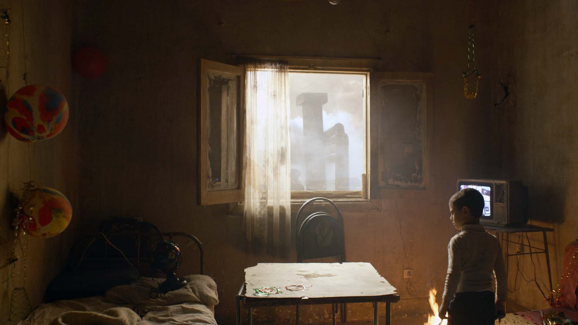 L'image montre une pièce sombre et légèrement en désordre, avec un enfant tourné vers une fenêtre ouverte. La lumière pénètre par cette fenêtre, révélant une scène extérieure brumeuse. À l'intérieur, on voit un lit, une table en bois et un vieux téléviseur. Des décorations festives, comme des ballons et des guirlandes, ajoutent une touche de couleur, bien que l'atmosphère générale semble mélancolique.