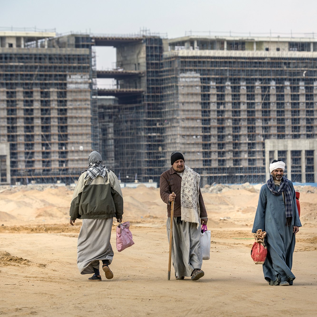 L'image montre un paysage de construction, avec plusieurs bâtiments inachevés en arrière-plan. Au premier plan, on voit quatre personnes marchant sur un sol de terre. Elles portent des vêtements traditionnels et semblent transporter des sacs. L'environnement est désertique, avec des matériaux de construction visibles autour. Le ciel est nuageux, contribuant à l'atmosphère de travail en cours.