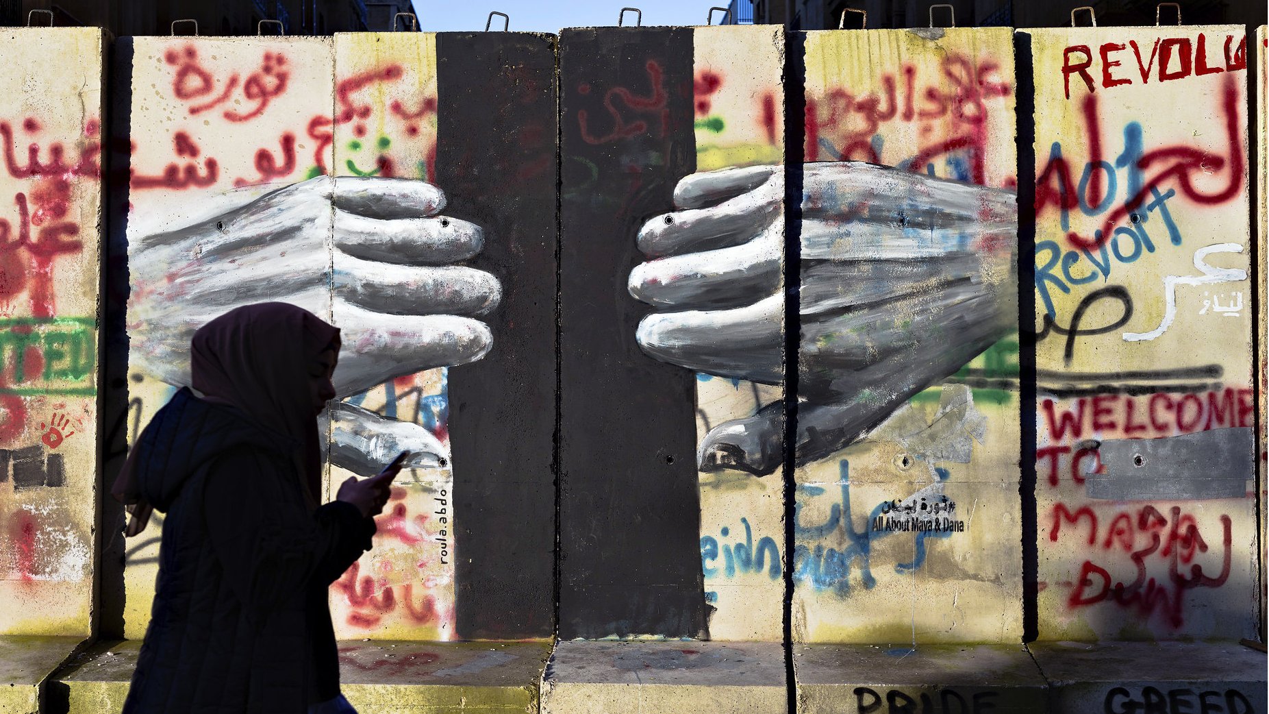 L'image montre un mur avec des graffitis colorés, dont le mot "Révolte" et d'autres messages en arabe et en anglais. Au centre, on peut voir des mains peintes qui semblent se toucher à travers une fissure du mur. Une silhouette d'une personne portant un hijab marche devant ce mur, avec un téléphone à la main. L'ensemble évoque des thèmes de résistance et de lutte sociale.