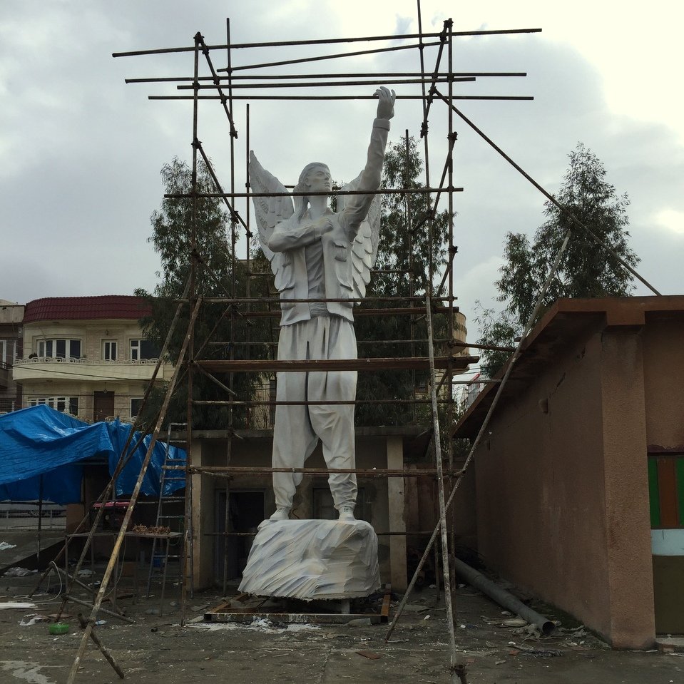 L'image montre une statue de grande taille, représentant un personnage avec des ailes, qui semble être en train d'être rénovée ou construite. Des échafaudages entourent la statue, indiquant qu'elle est encore en travaux. En arrière-plan, on peut entrevoir des bâtiments et une structure en toile bleue, suggérant une ambiance urbaine. Le ciel est nuageux, ce qui donne une atmosphère un peu mélancolique à la scène.