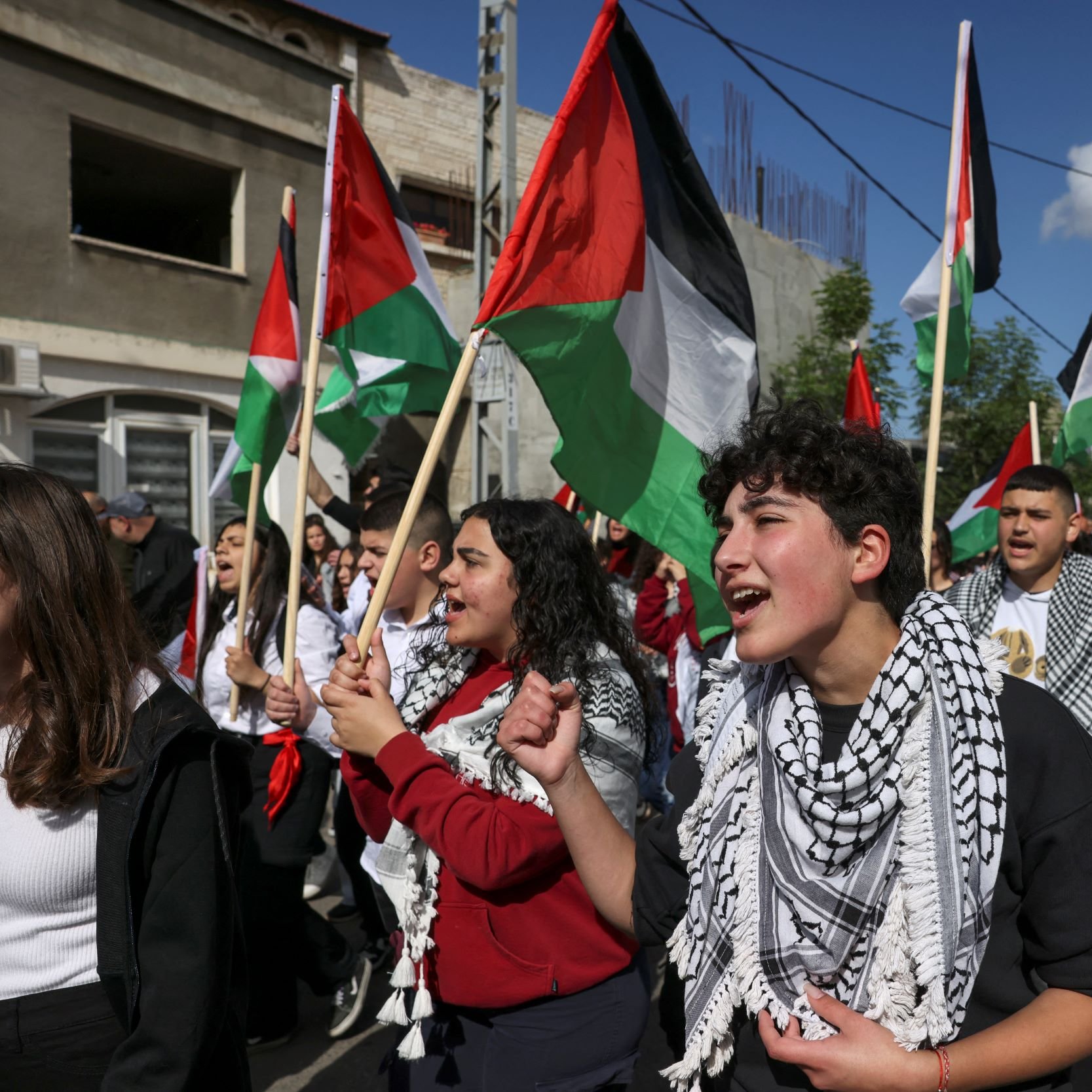 L'immagine mostra un gruppo di persone che partecipano a una manifestazione. Molti manifestanti tengono in mano bandiere palestinesi, caratterizzate da strisce nere, bianche e verdi e da un triangolo rosso. Alcuni di loro indossano kefiah, tradizionali sciarpe palestinesi. Le espressioni sui loro volti trasmettono passione e determinazione mentre cantano o gridano slogan, contribuendo a creare un'atmosfera di unità e attivismo. Sullo sfondo, si intravedono edifici e il cielo è parzialmente nuvoloso.