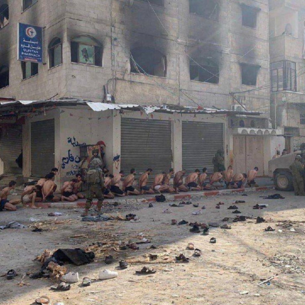 Immagine di un gruppo di persone su un marciapiede, sotto sorveglianza militare, in un contesto urbano.