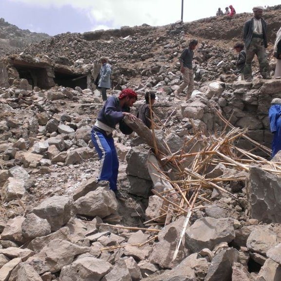 L'image montre un groupe de personnes travaillant dans les décombres d'un bâtiment effondré. Elles semblent s'affairer à dégager des pierres et des débris, avec des outils improvisés. L'environnement paraît montagneux et accidenté, avec des restes de structures anciennes en arrière-plan. L'atmosphère est celle de l'effort collectif dans un contexte de reconstruction ou d'aide humanitaire.