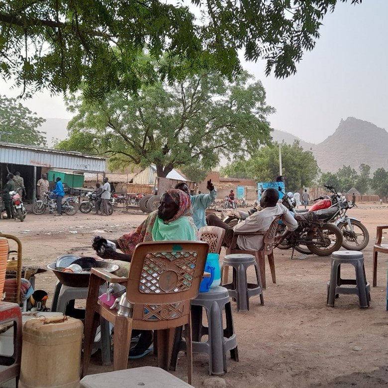 L'image montre une scène de la vie quotidienne dans un village. On y voit plusieurs chaises de différents styles et couleurs, dont certaines sont empilées. À l'arrière-plan, il y a des maisons, des collines, et plusieurs motos. La végétation est présente avec quelques arbres, offrant de l'ombre. On aperçoit également des personnes assises et se déplaçant, ce qui donne un aperçu de l'ambiance locale. Les routes sont poussiéreuses, suggérant une atmosphère rurale.