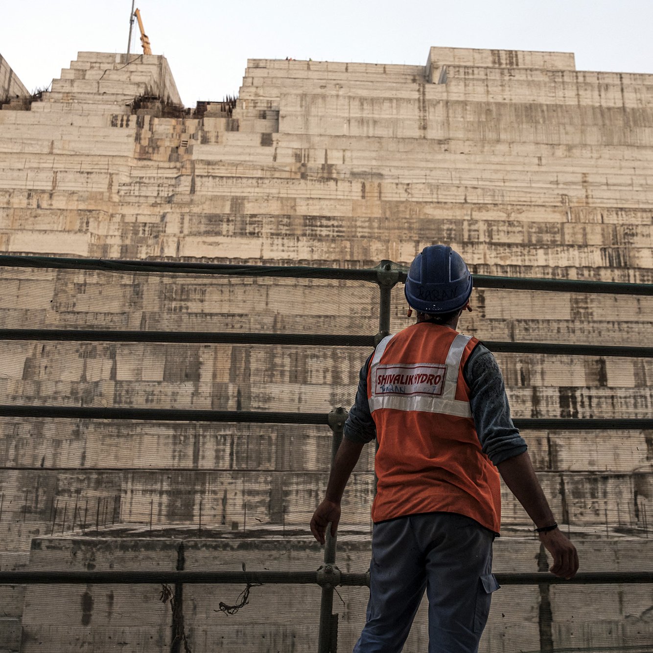 تظهر في الصورة عامل بناء يرتدي سترة واقية زاهية اللون وخوذة، وهو يقف أمام هيكل كبير من الخرسانة يبدو أنه في مرحلة الإنشاء. خلفه، تُلاحظ طبقات من المواد البنائية، مما يشير إلى مشروع بناء ضخم. يظهر في الخلفية معدات البناء مثل رافعة، مما يضيف إلى جو العمل المكثف والحركة في المكان. السماء تميل إلى اللون الأزرق، مما يوحي بأن الوقت هو إما الصباح الباكر أو بعد الظهر.