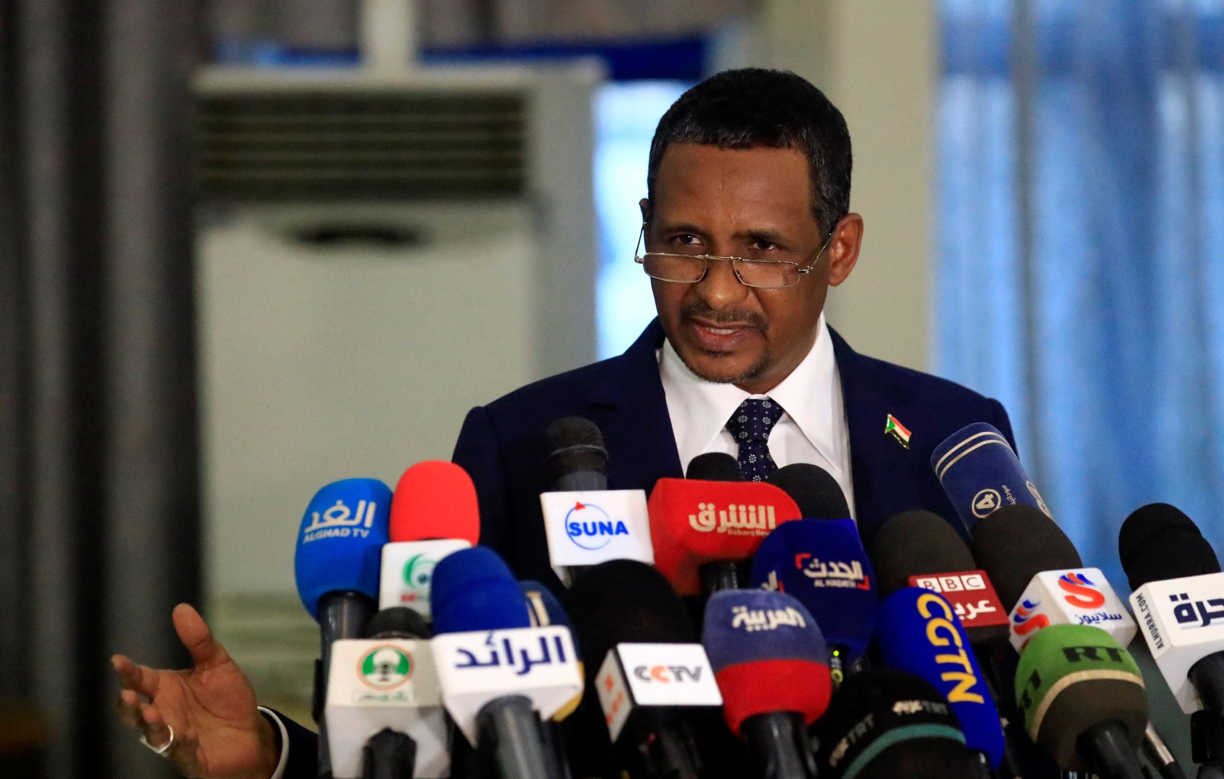 در این تصویر، مردی در حال صحبت کردن در جلوی میکروفن‌های زیادی است. او لباس رسمی به تن دارد و به نظر می‌رسد در حال ارائه یک سخنرانی یا بیانیه است. در پس‌زمینه، تعدادی دوربین و میکروفون وجود دارد که نشان‌دهنده جوی رسانه‌ای و توجه عمومی به صحبت‌های اوست. فضا به‌نظر رسمی و جدی می‌رسد.