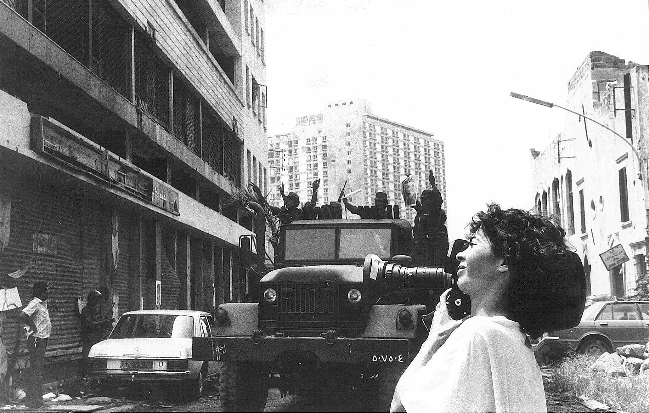 L'image montre une scène urbaine où une femme tient un appareil photo, prenant probablement des clichés d'une situation troublée. En arrière-plan, on peut voir un véhicule militaire sur une route, avec plusieurs personnes en uniforme visible sur le camion. Les bâtiments autour semblent en mauvais état, avec des stores métalliques fermés. L'atmosphère dégage une impression de tension ou de conflit. La femme, avec des cheveux bouclés, semble concentrée sur sa prise de vue, capturant un moment significatif dans un contexte difficile.