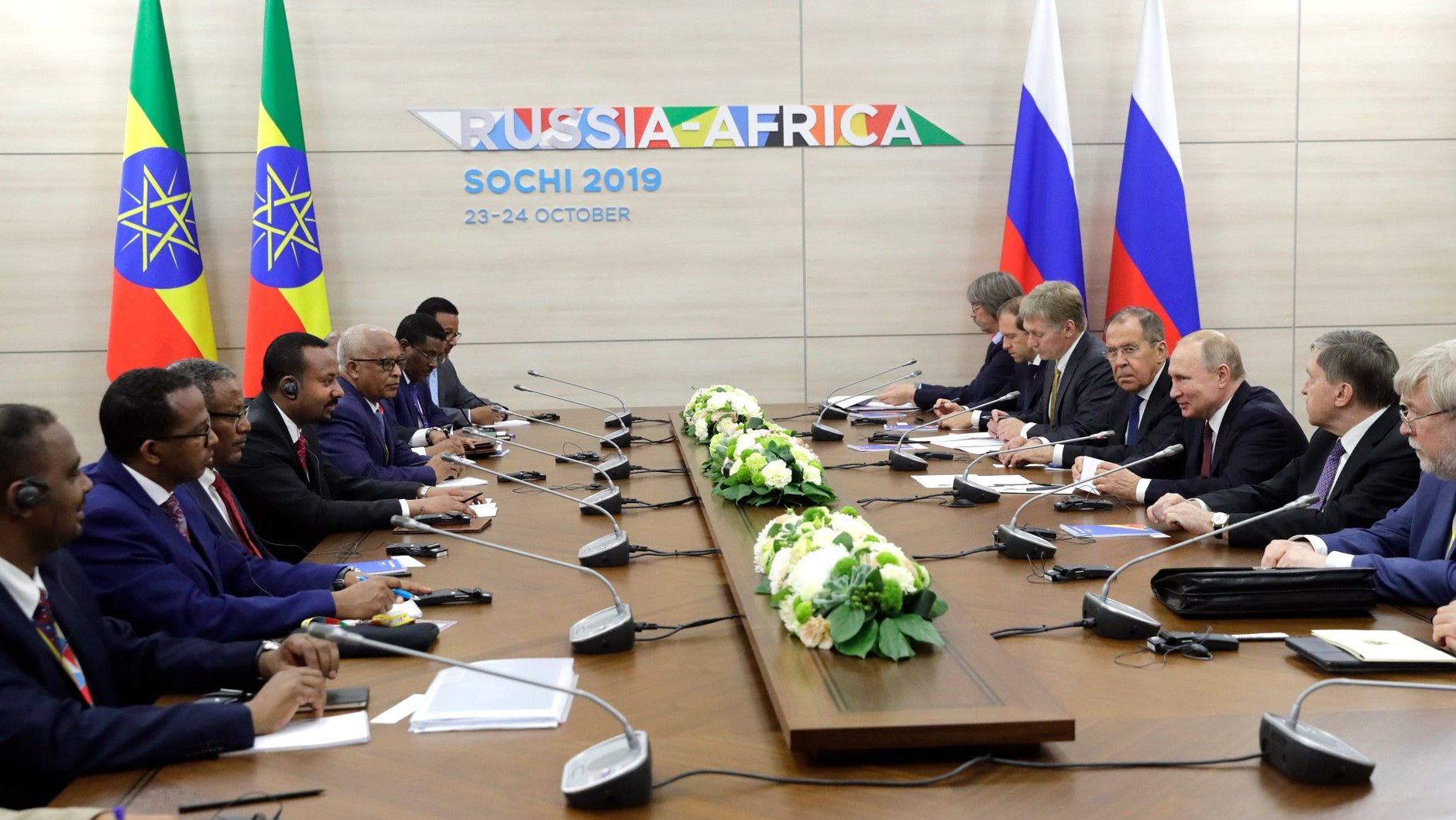 L'image montre une réunion officielle tenue à Sochi, en Russie, en octobre 2019, dans le cadre d'un sommet Russie-Afrique. On peut voir des représentants de plusieurs pays africains, ainsi que des dignitaires russes, assis autour d'une grande table de conférence. Des drapeaux représentant l'Éthiopie et un autre pays africain sont visibles en arrière-plan. Au centre de la table, il y a des arrangements floraux. Les participants semblent engagés dans une discussion, avec certains utilisant des écouteurs pour la traduction.